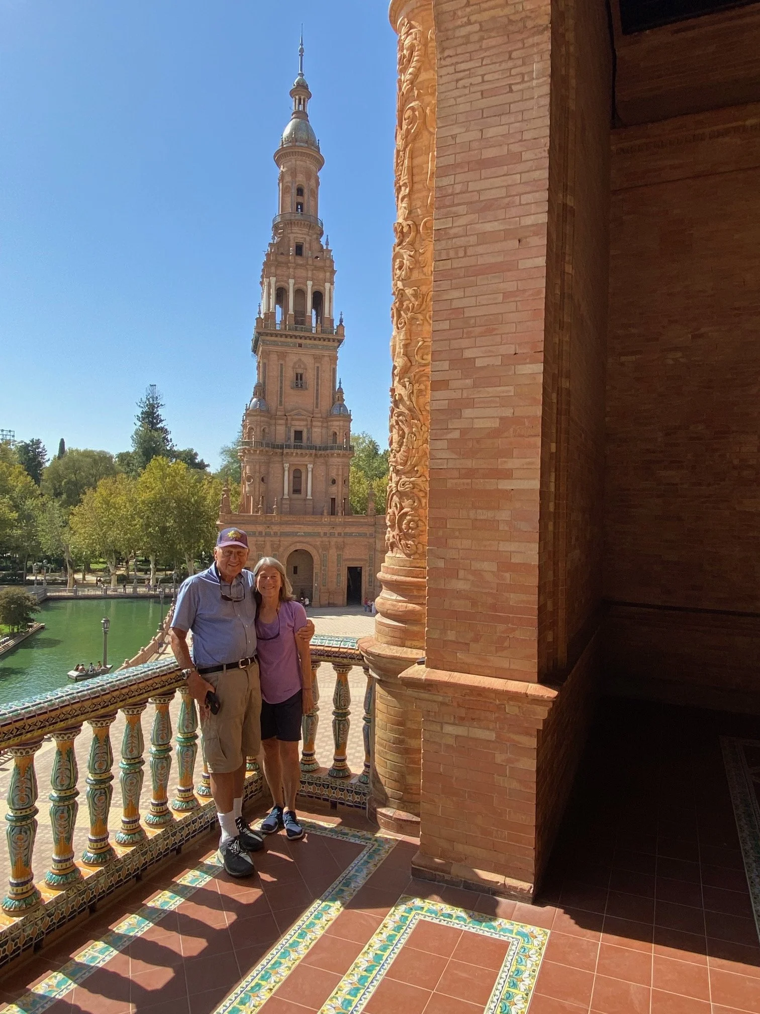 A smiling man and woman standing together on a decorated balcony with colorful tiles, with a large historic tower and lush green trees in the background on a sunny day.