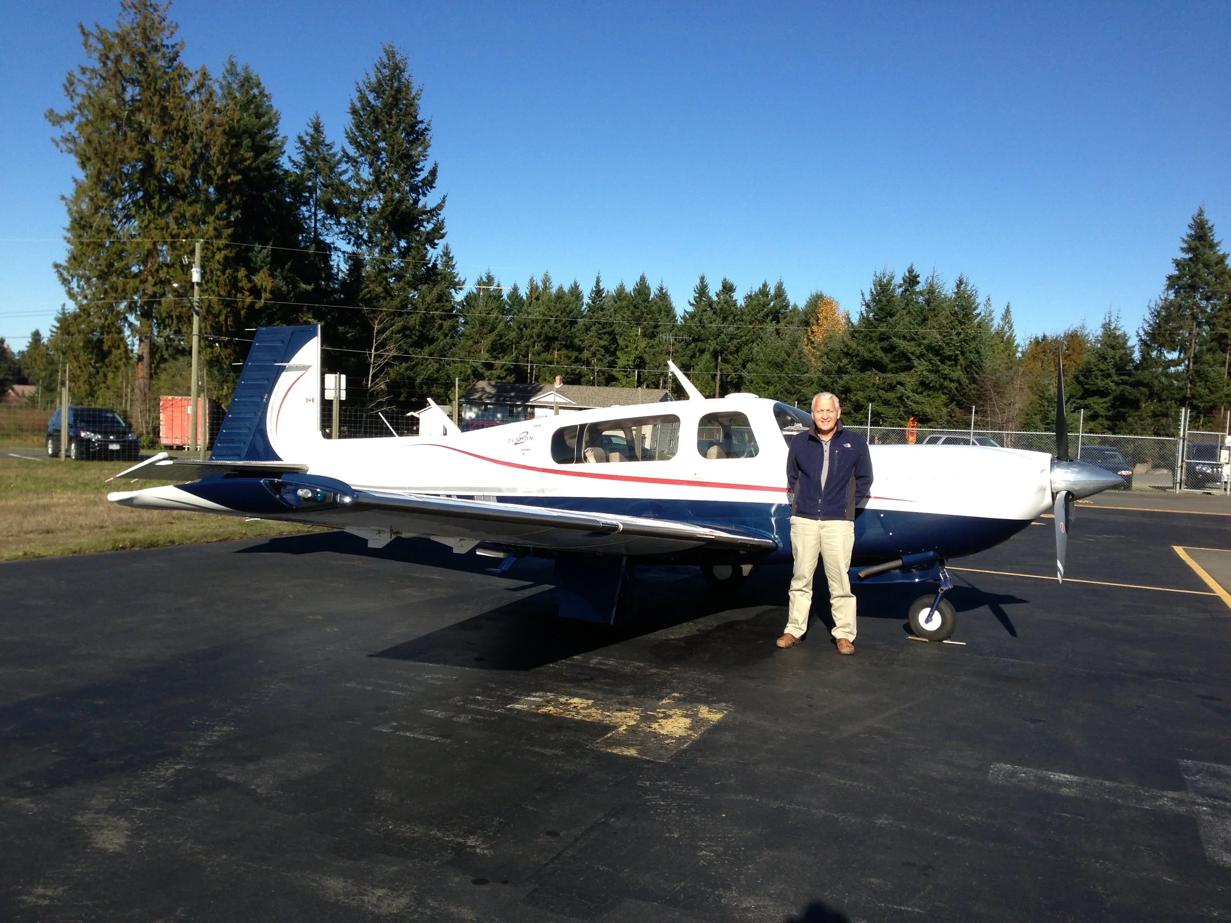 A man stands next to a white and blue small airplane parked on a tarmac, with trees and houses in the background during daytime.
