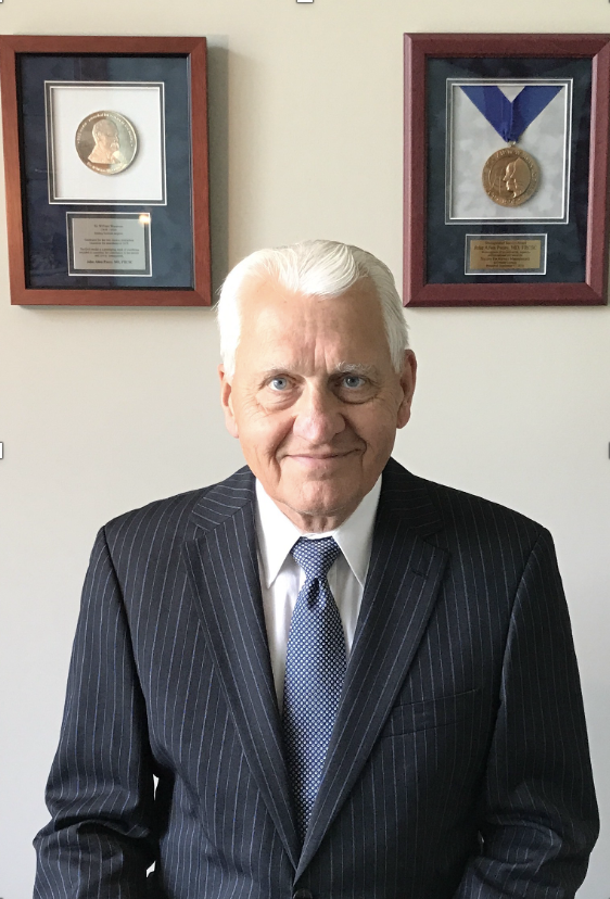 A senior man in a pinstripe suit and tie sitting in front of a white wall with two framed medals, one with a profile of a person and the other with a ribbon, hanging behind him.