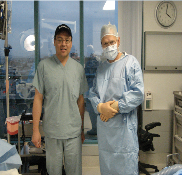 Two medical professionals standing in a hospital operating room, one wearing scrubs and a cap, the other in surgical attire with a mask and gloves.