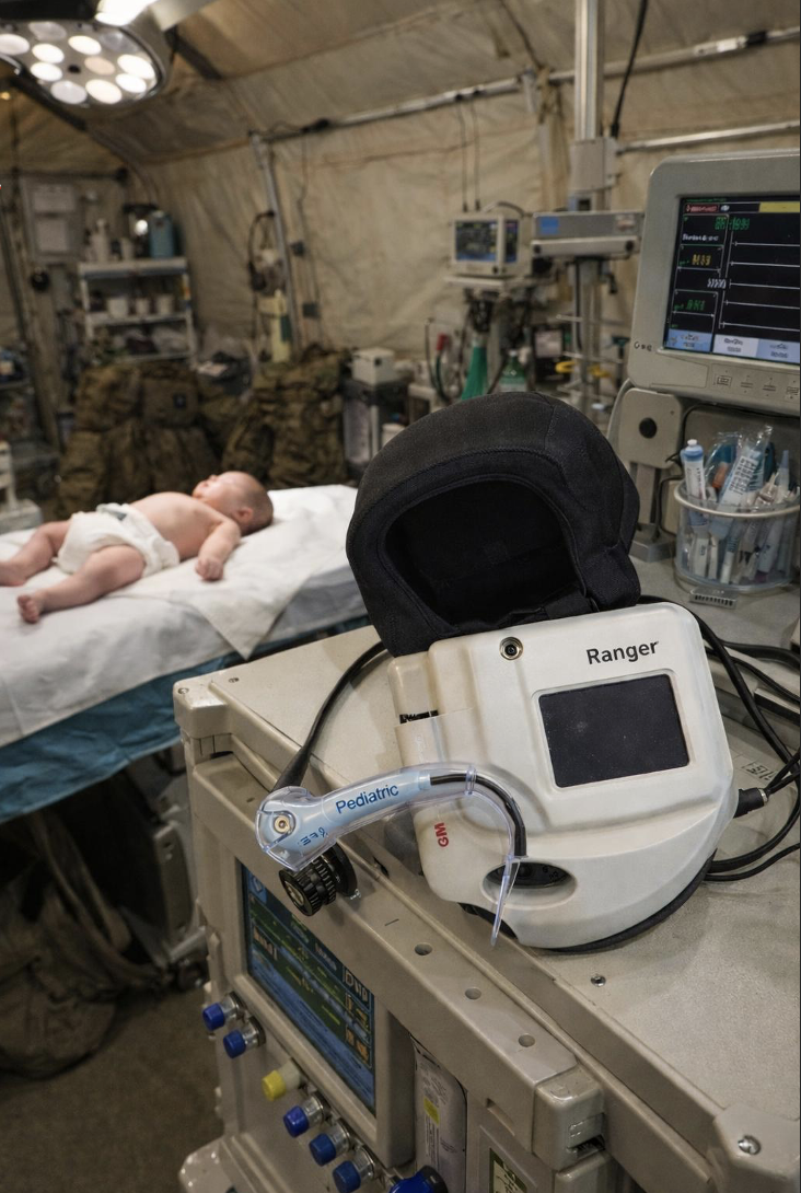Inside a military tent decorated as a makeshift emergency room, a newborn baby lies on a hospital bed with medical equipment and monitors around.