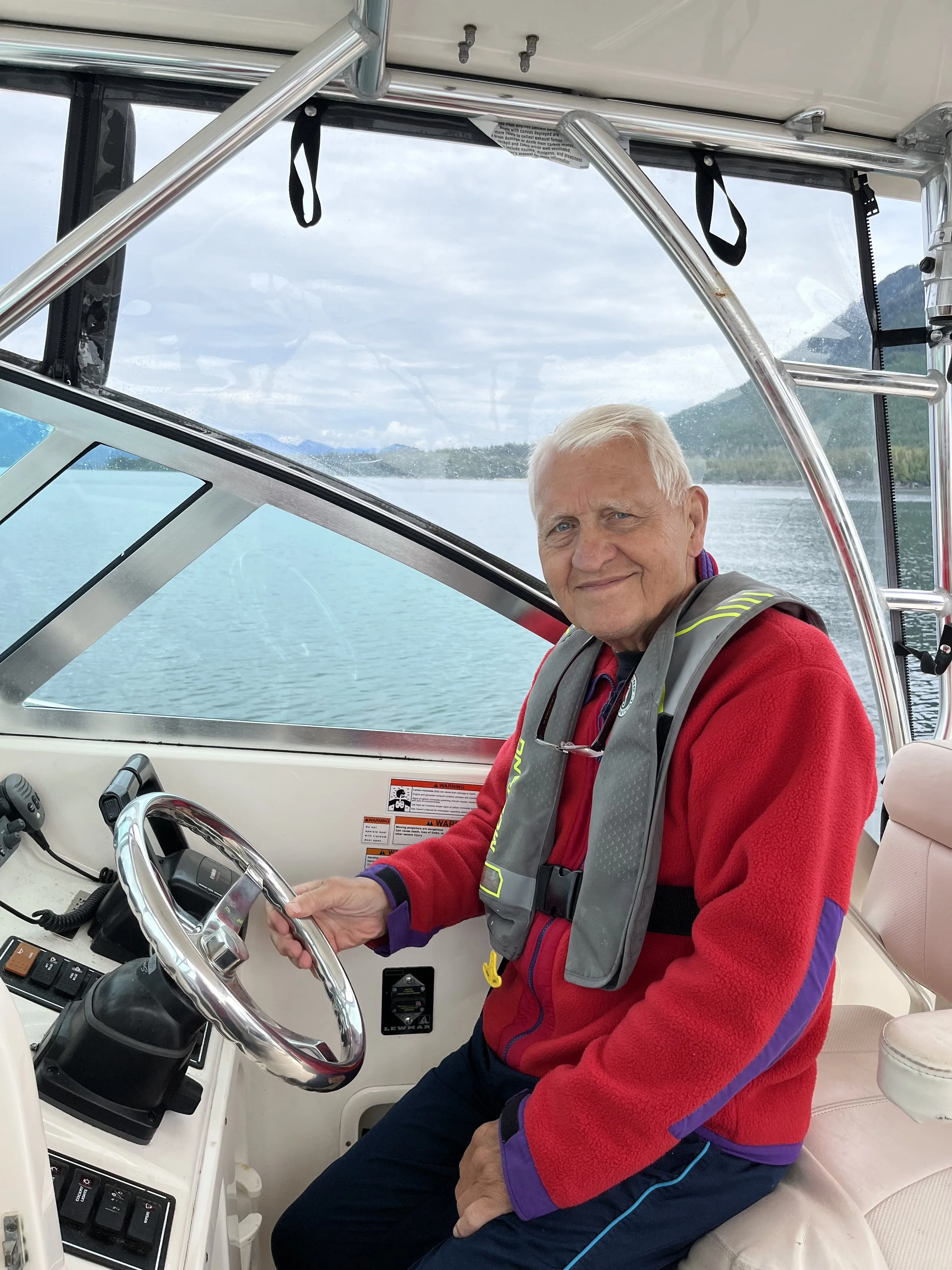 An older man with short white hair sitting at the helm of a boat, holding the steering wheel, with water and mountains in the background outside the boat's windows.