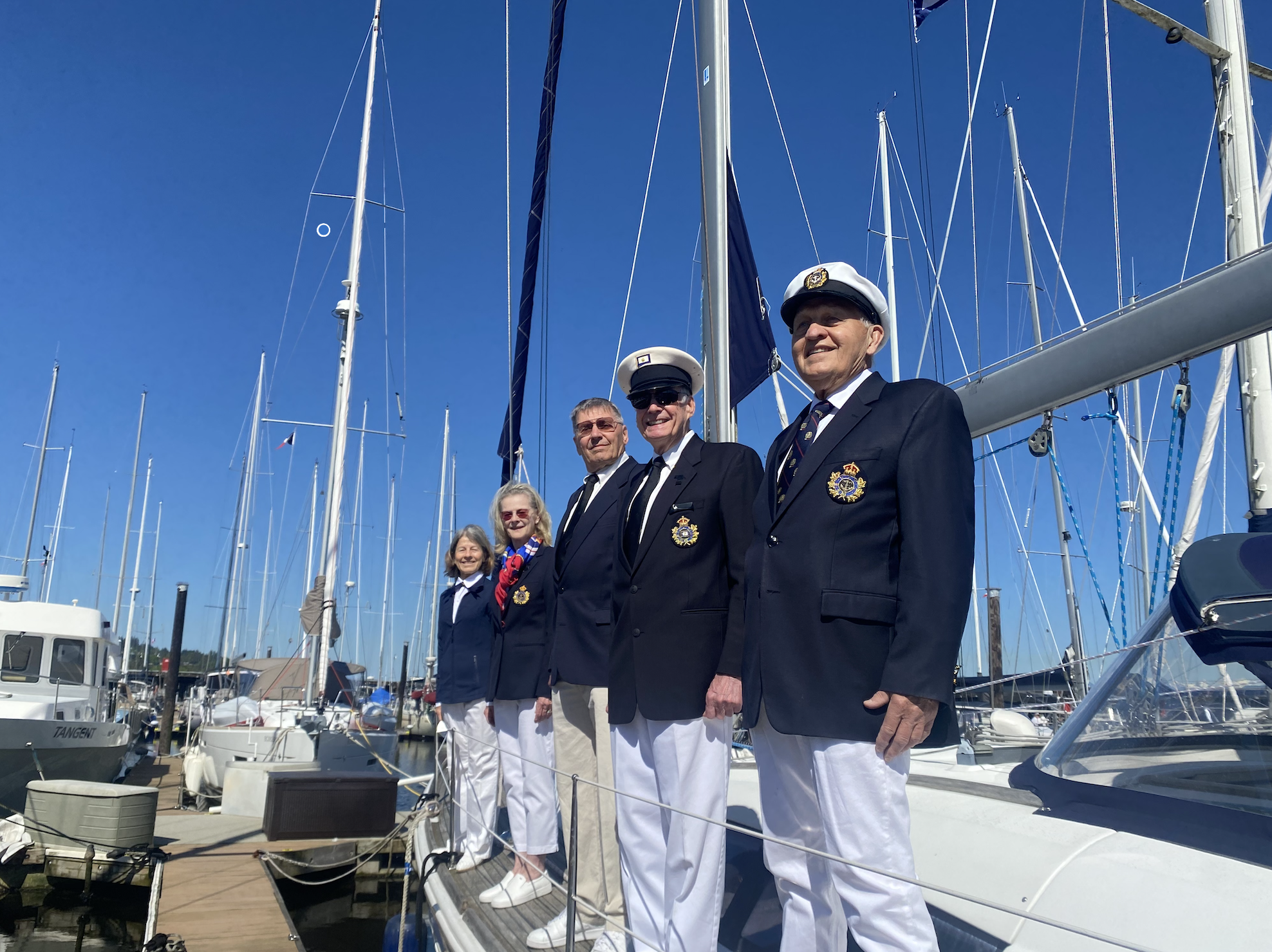 Group of people, including men in navy uniforms with hats and women in casual clothing, standing on a dock at a marina with sailboats and masts in the background.