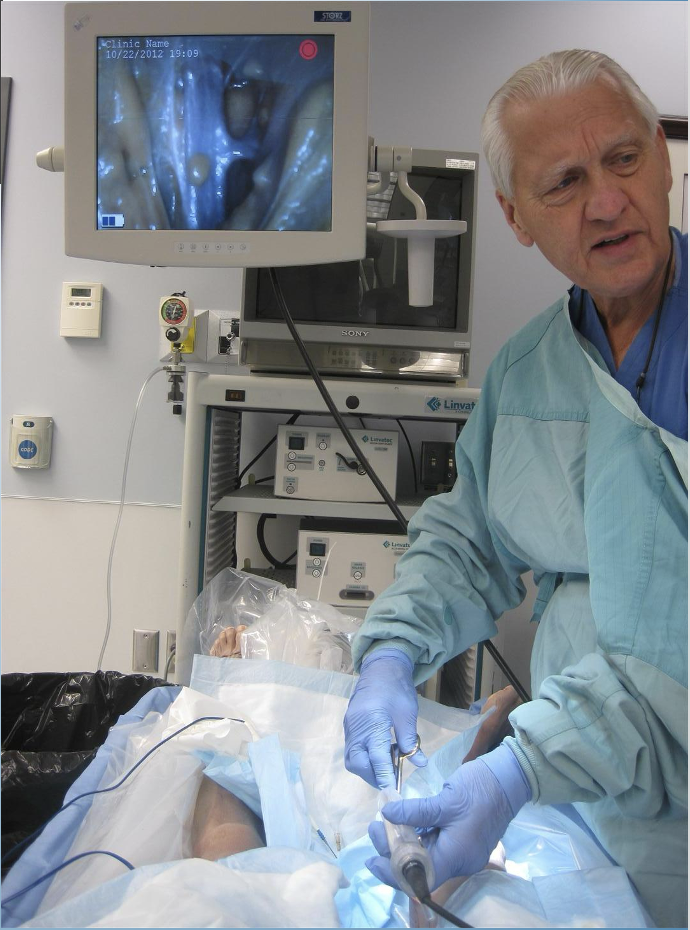 A doctor in surgical gown and gloves is performing a medical procedure on a patient in an operating room. Medical equipment, monitors, and screens displaying medical images are visible.