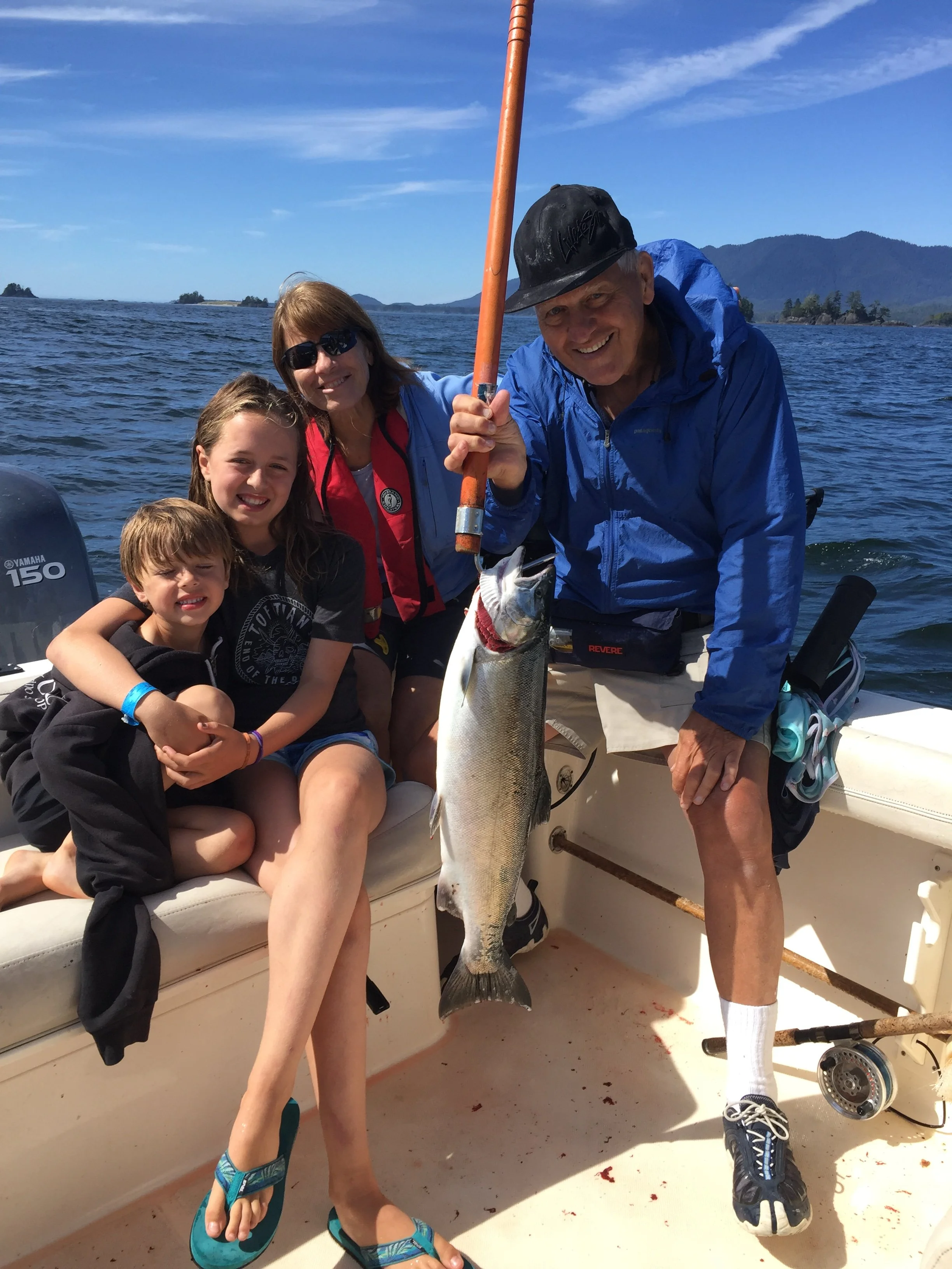 Four people on a boat holding a large fish they caught, with water and mountains in the background.