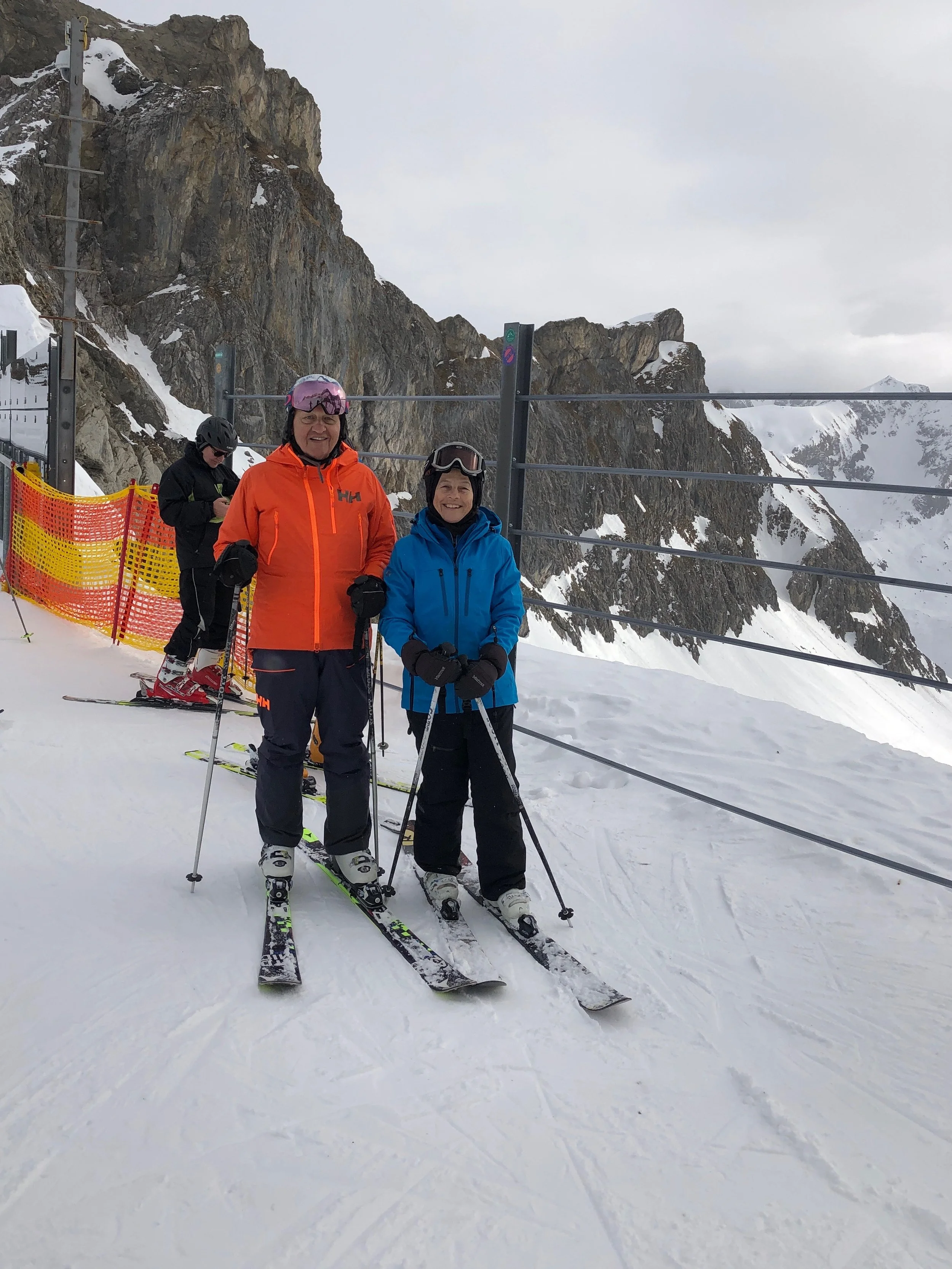 Two people standing on a snowy mountain with ski gear, smiling, with rocky mountain cliffs in the background.
