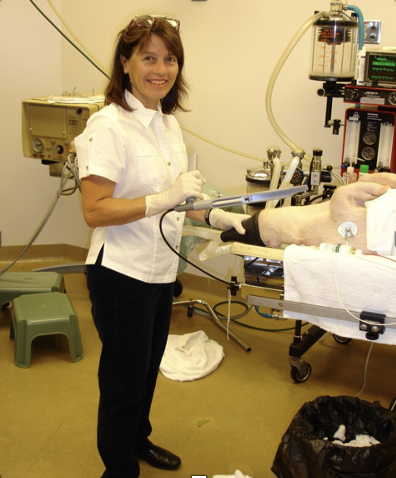 A female nurse or technician in a medical setting, smiling with medical equipment and a patient lying on a hospital bed.