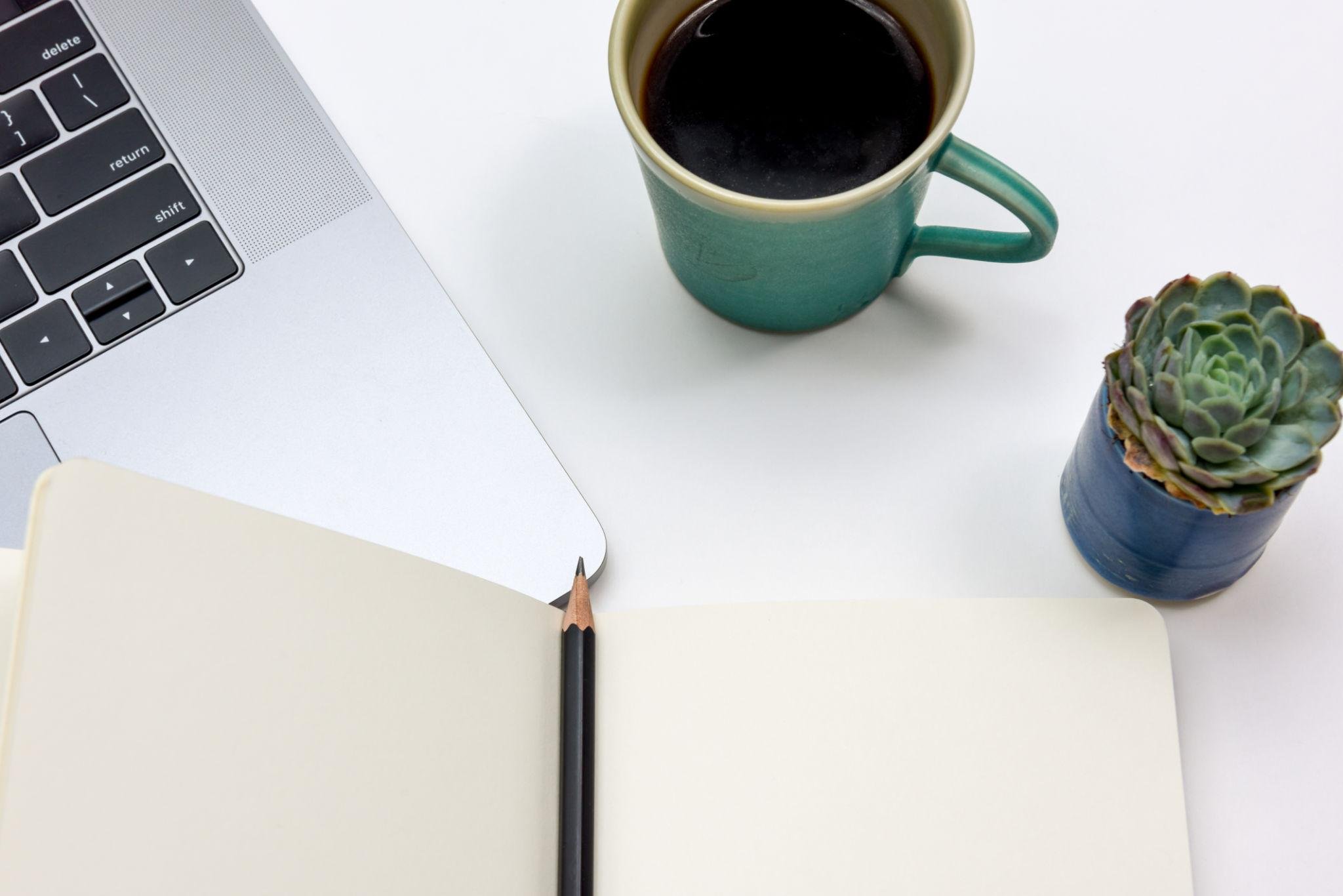 Top view of a workspace with an open notebook, a pencil, a laptop keyboard, a mug of black coffee, and a potted succulent plant on a white surface.