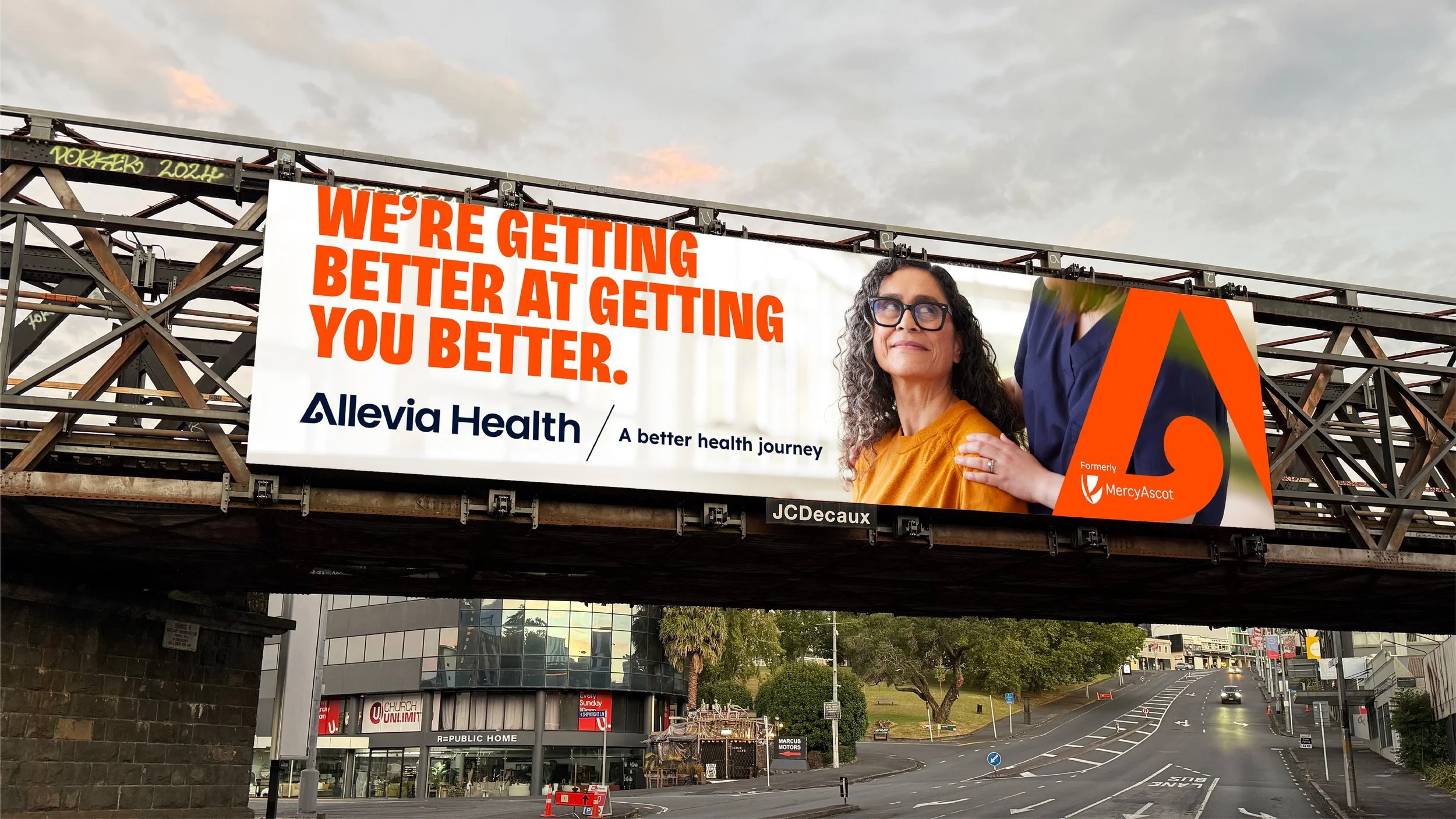 An outdoor billboard with a woman and a healthcare message. The billboard reads "We're getting better at getting you better." It is promoted by Allevia Health, formerly MercyAscot. The woman has curly gray hair, wears glasses, and is dressed in an orange shirt. She appears to be smiling and looking upwards. The billboard is mounted on a highway bridge, with trees and a street below.