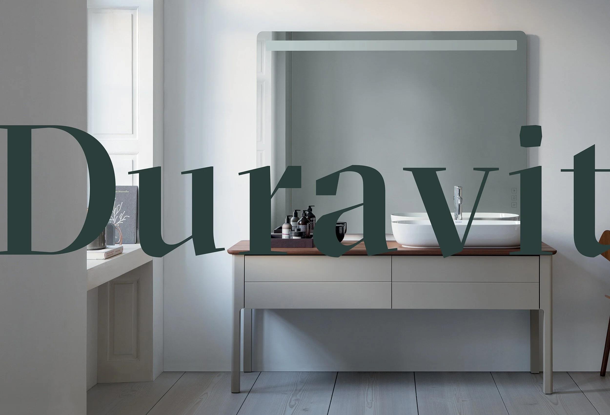 A modern bathroom with a white sink on a wooden countertop, bottles of toiletries, and a large mirror with a light gray frame on the wall.