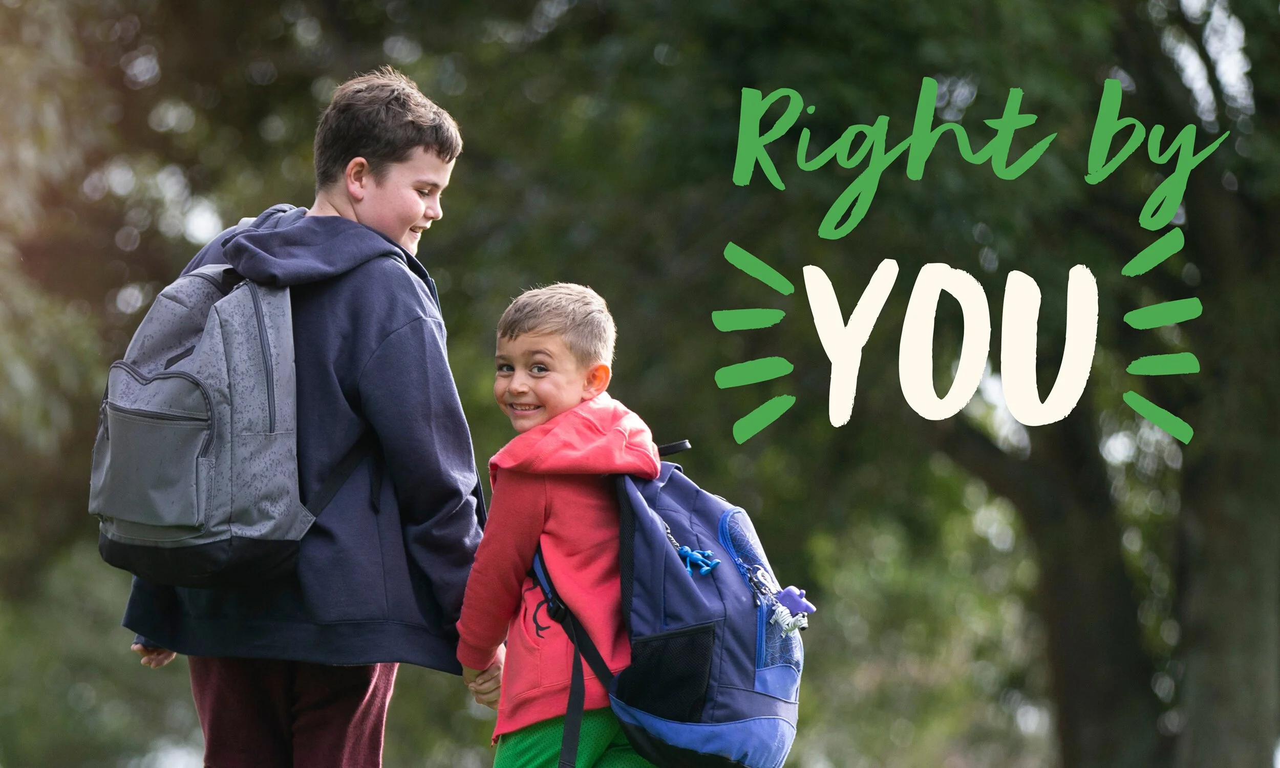 Two boys with backpacks walking outdoors, holding hands, and smiling, with the text 'Right by YOU' in green and white over a blurred green background.