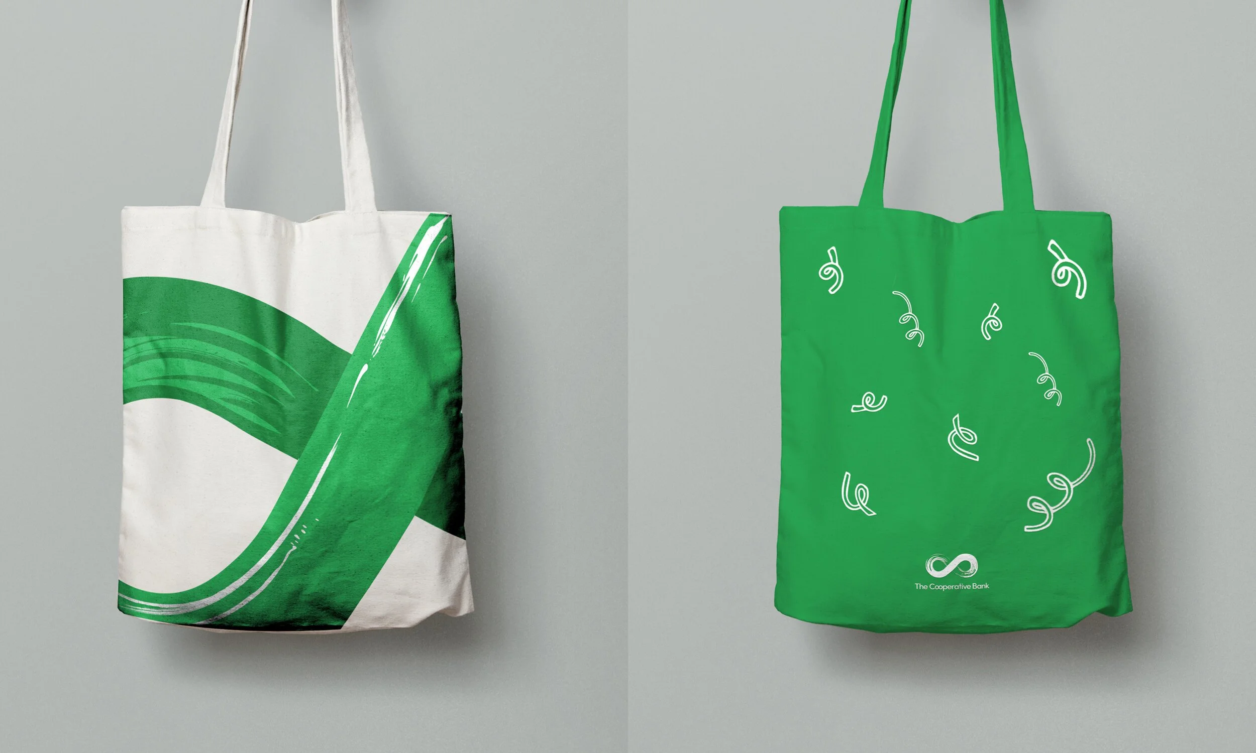 Two reusable tote bags hanging against a plain gray background, one white with green abstract design, and one green with white confetti-like swirls, both featuring the logo of The Cooperative Bank.