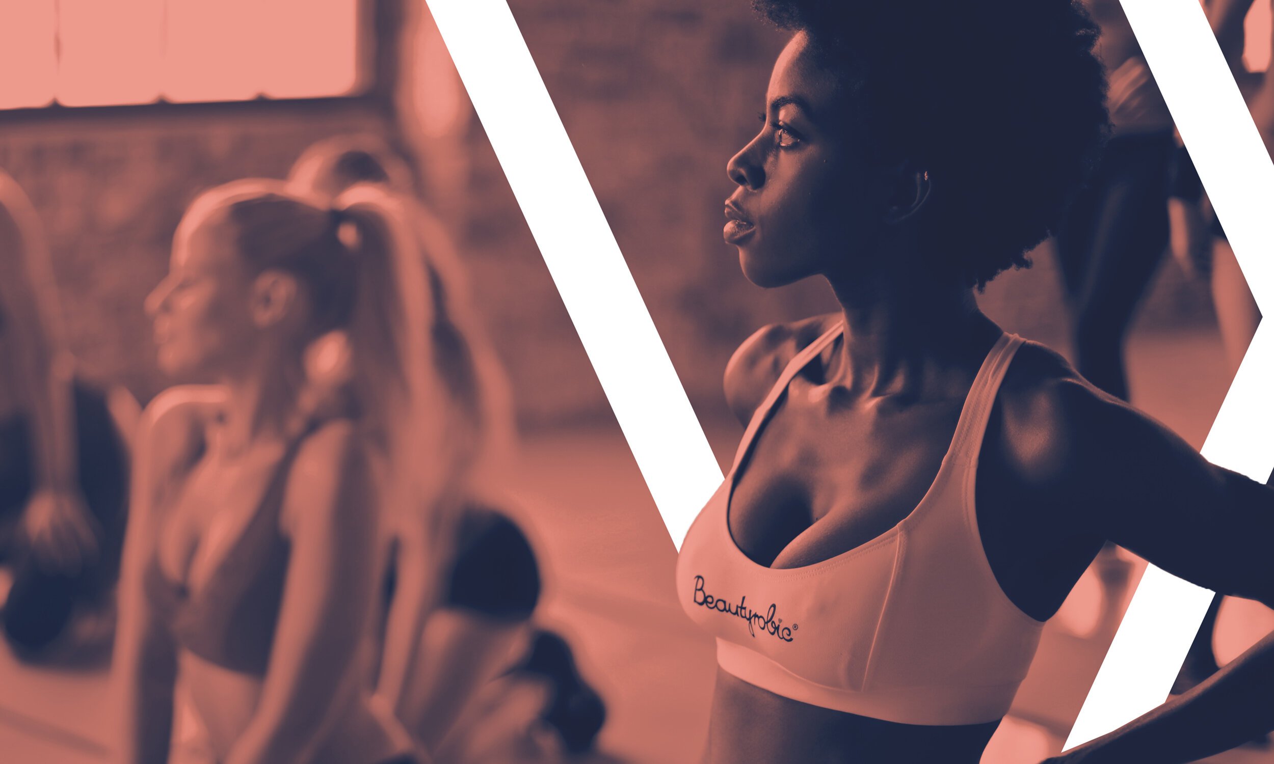 A woman standing with her hands on her hips, with her head turned to the side, during a workout session at a gym. She is wearing a sports bra with the words 'Beautylogi' on it, and her hair is styled in a natural, voluminous afro.