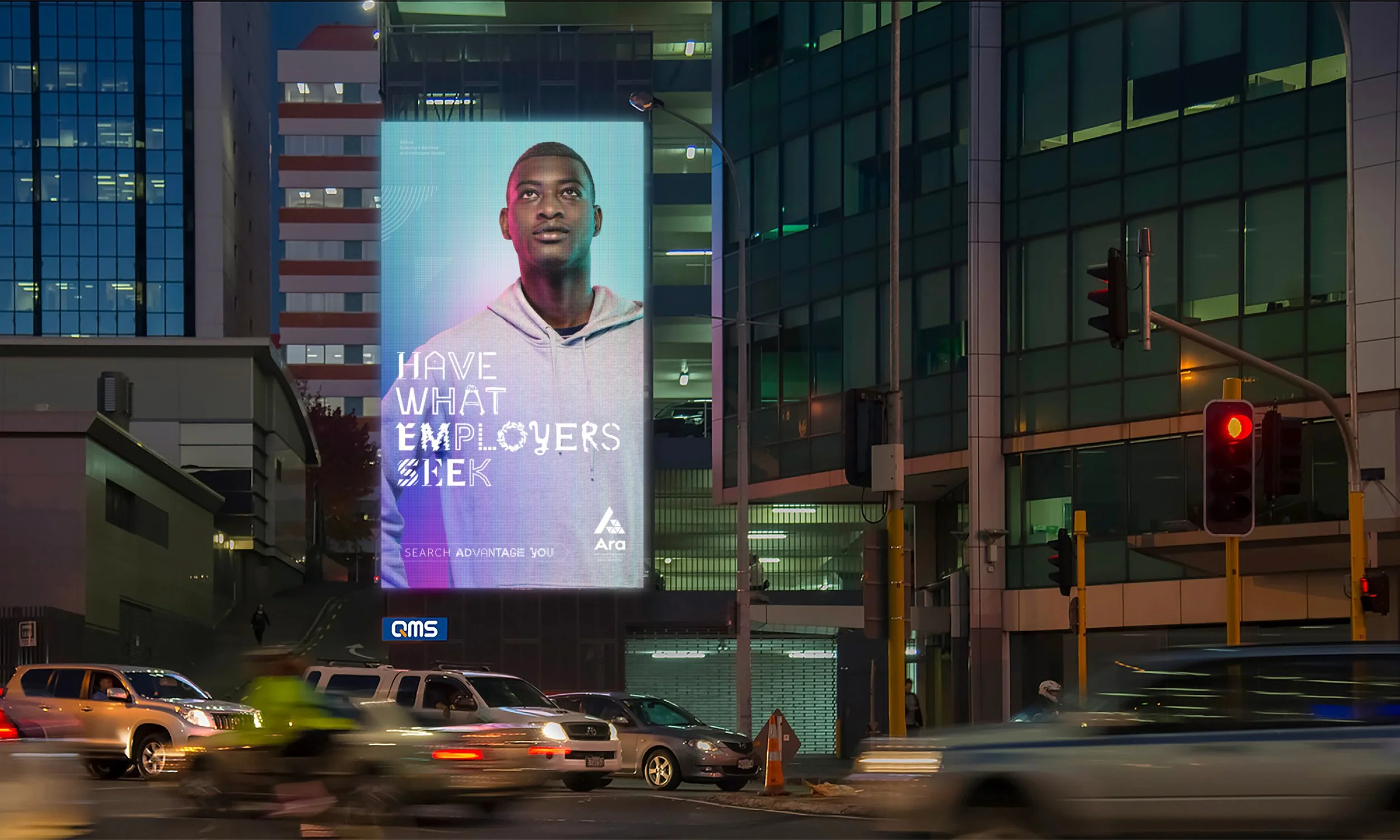 A large digital billboard in an urban area displaying a portrait of a young man with the message 'Have What Employers Seek' and the logo 'Ara.' Surrounding the billboard are modern glass buildings, traffic lights, and vehicles on the street, indicating busy city traffic at dusk or nighttime.