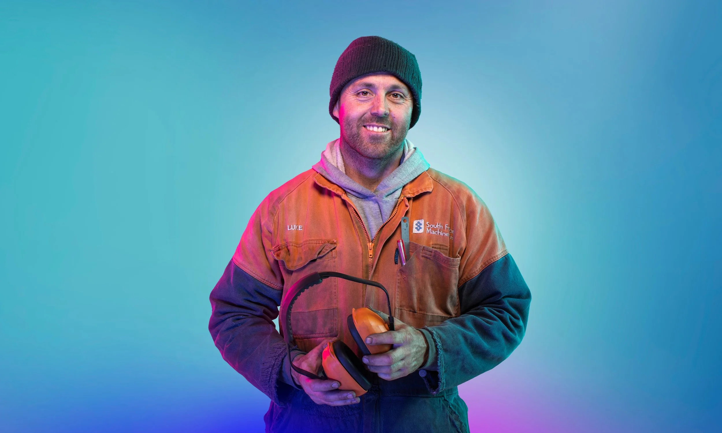 A man wearing a black beanie, gray hoodie, and a brown jacket with a name tag that says 'LUKE' and 'South Free Machine,' is smiling and holding a power tool, standing against a colorful gradient background.