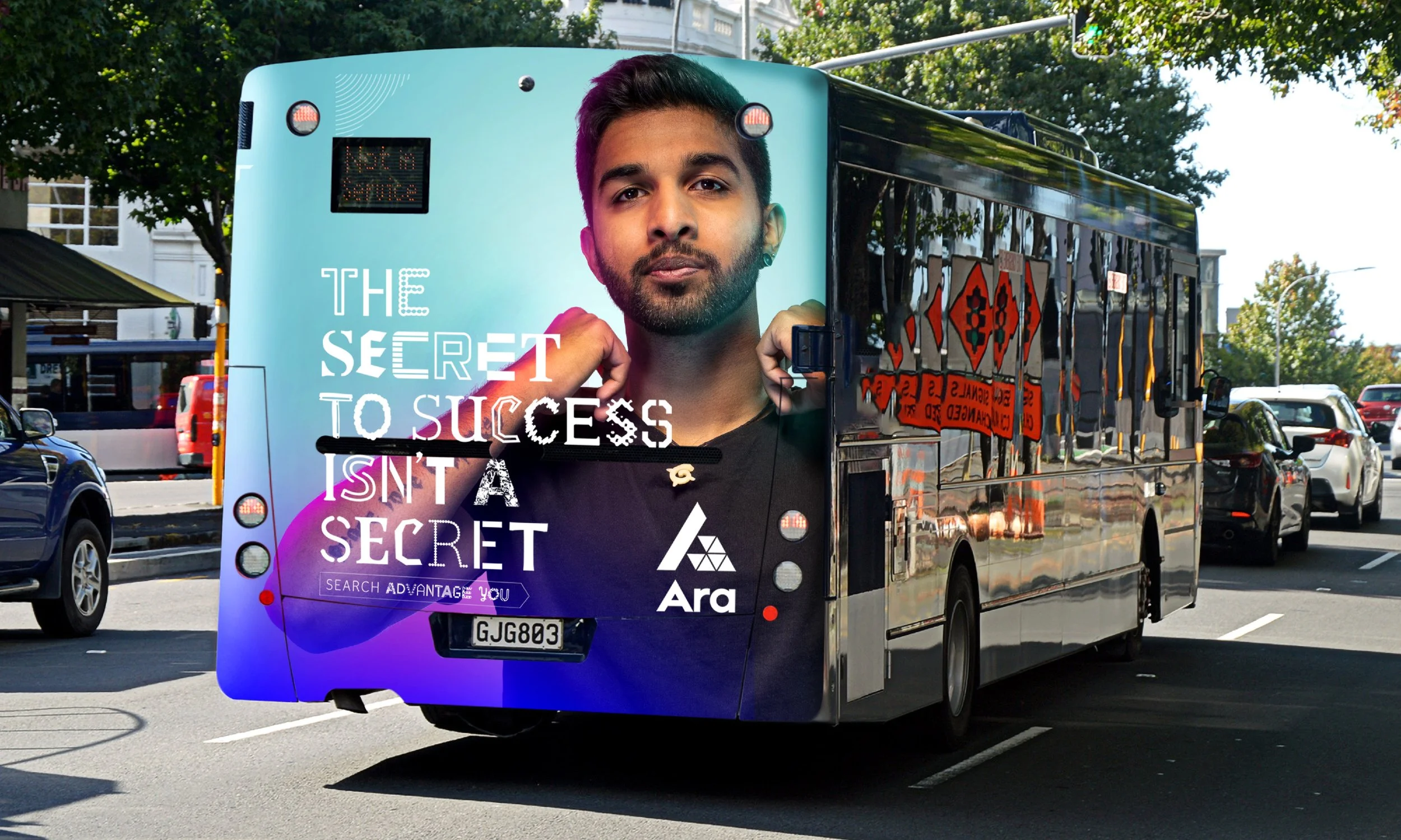 A city bus with a colorful advertisement of a young man with dark hair and a beard, partially showing the words 'The Secret to Success Isn't a Secret' and the logo 'Ara' on the back of the bus, driving on a street with parked cars and trees.