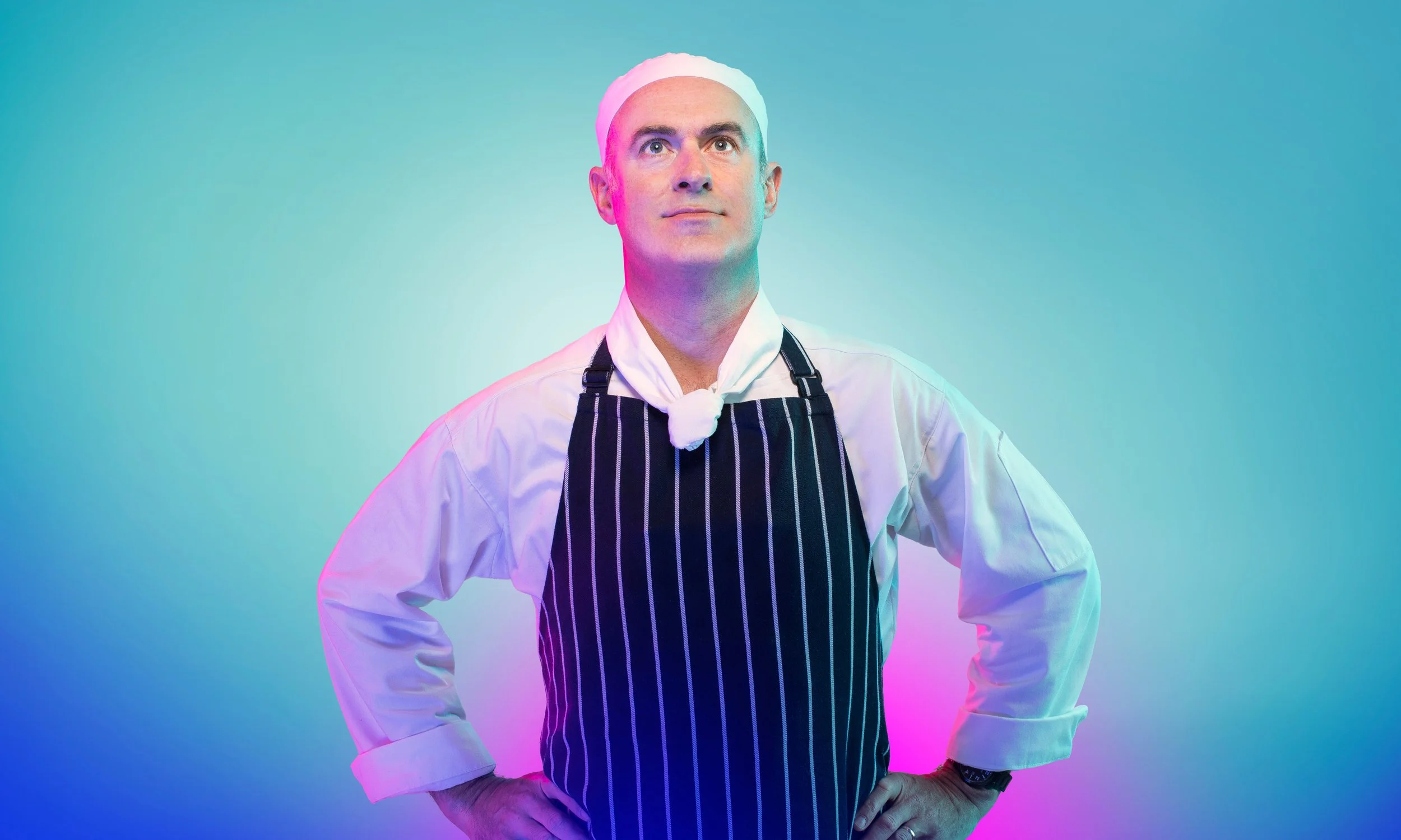A male chef wearing a white chef hat, white shirt, and black apron with white stripes, standing with hands on hips against a colorful gradient background.