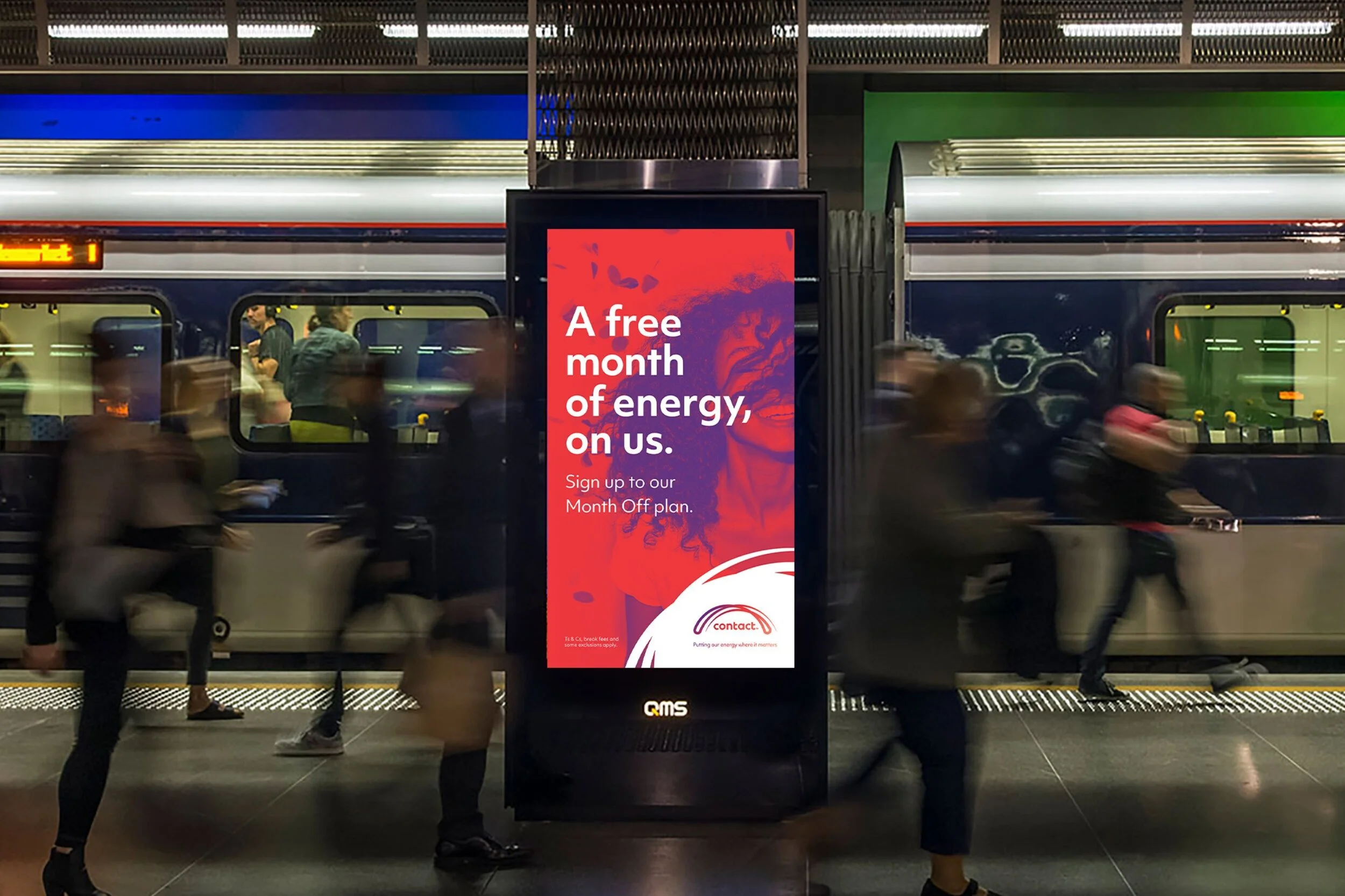A train station platform with a digital advertisement board in the center that displays a red and purple ad reading 'A free month of energy, on us.' People are walking past the ad, and the train behind has a blue and green color scheme with windows showing the interior and passengers.