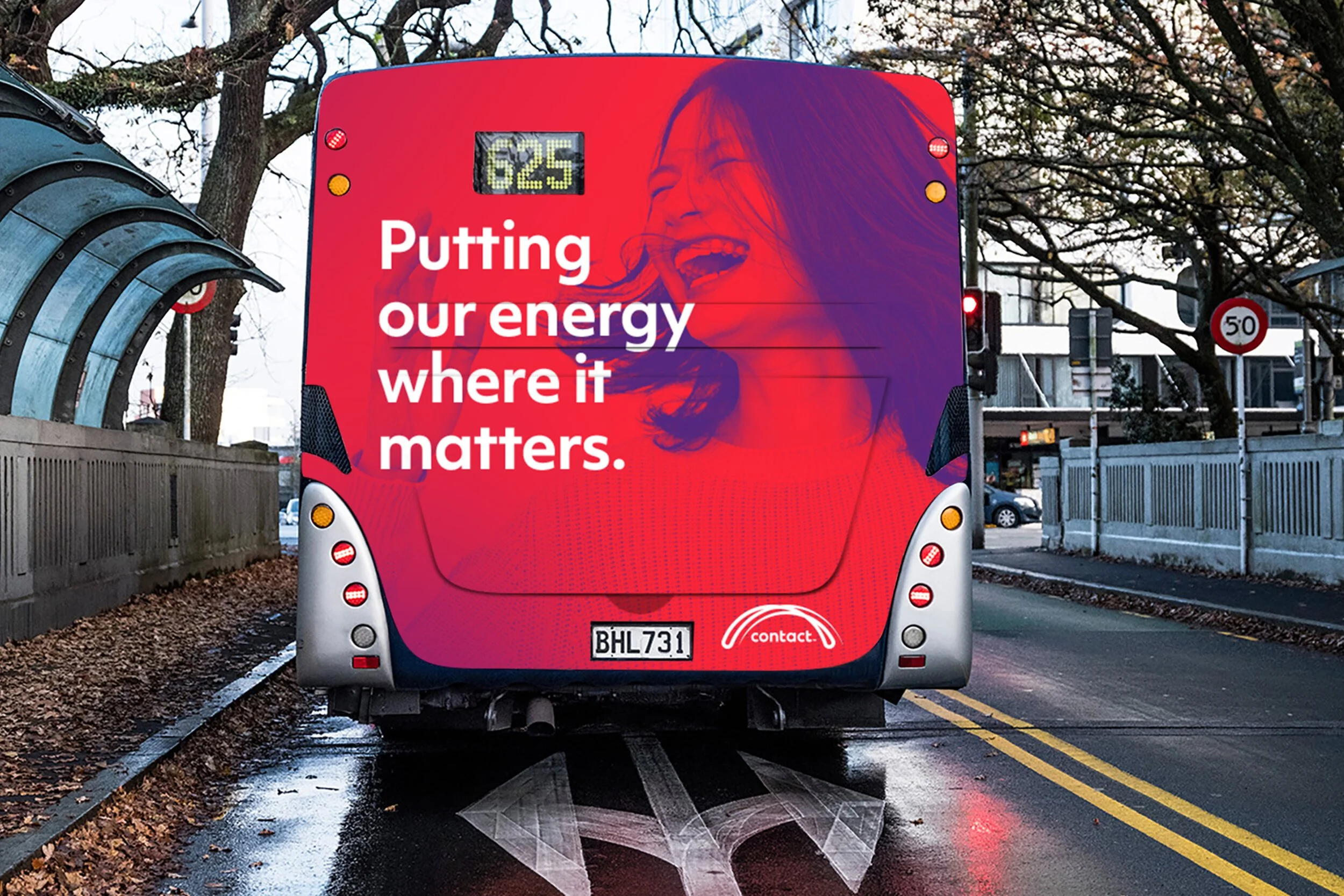 The back of a red bus displaying a digital screen with the number 625. The bus has a colorful advertisement featuring a woman laughing, with the text 'Putting our energy where it matters.' The bus's license plate reads BHL731 and it has contact information. The bus is stopped on a wet city street, with a cityscape and leafless trees in the background.