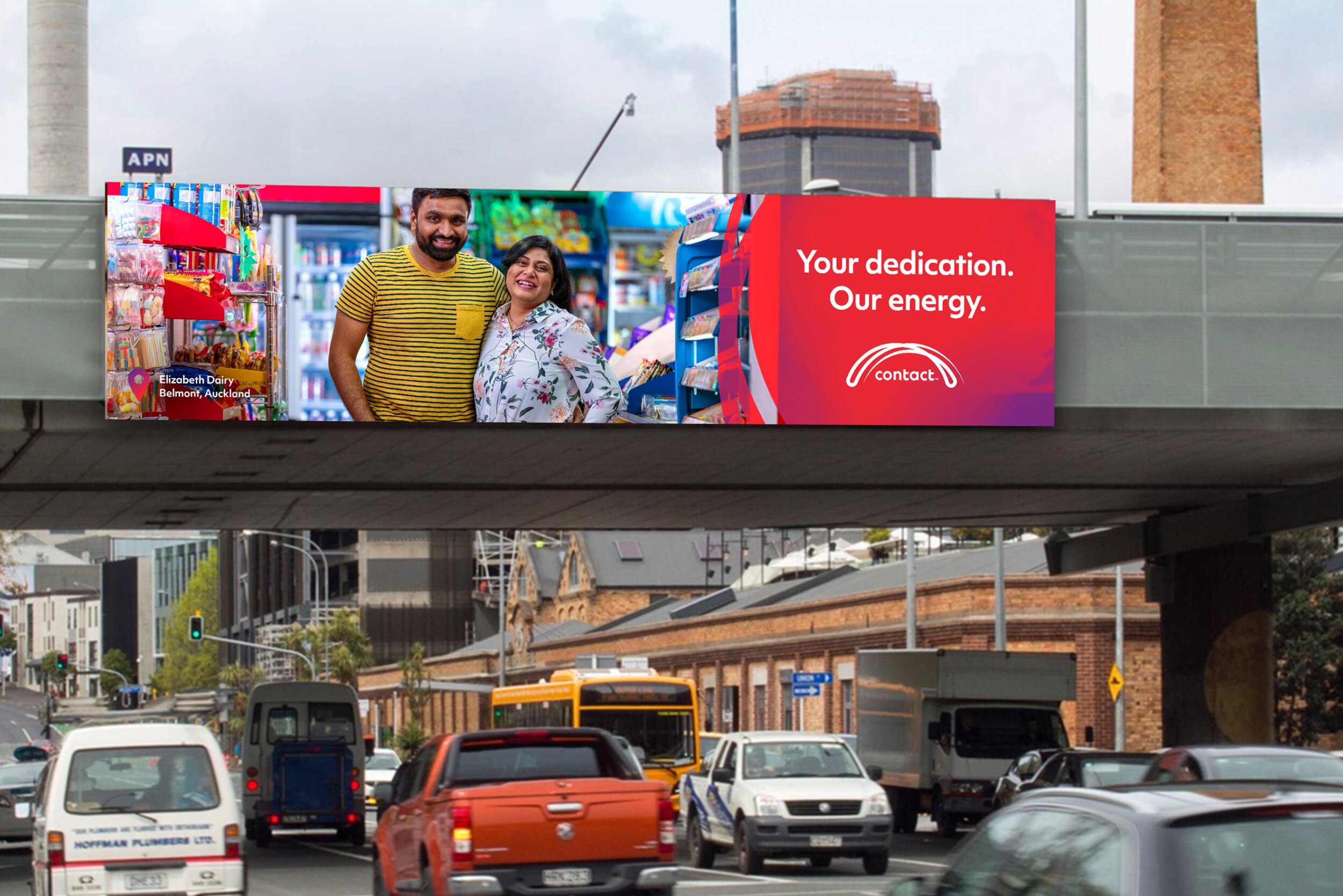 A large digital billboard on an overpass featuring a smiling couple inside a store, with the message 'Your dedication. Our energy.' and a logo, set against an urban background with traffic and buildings.