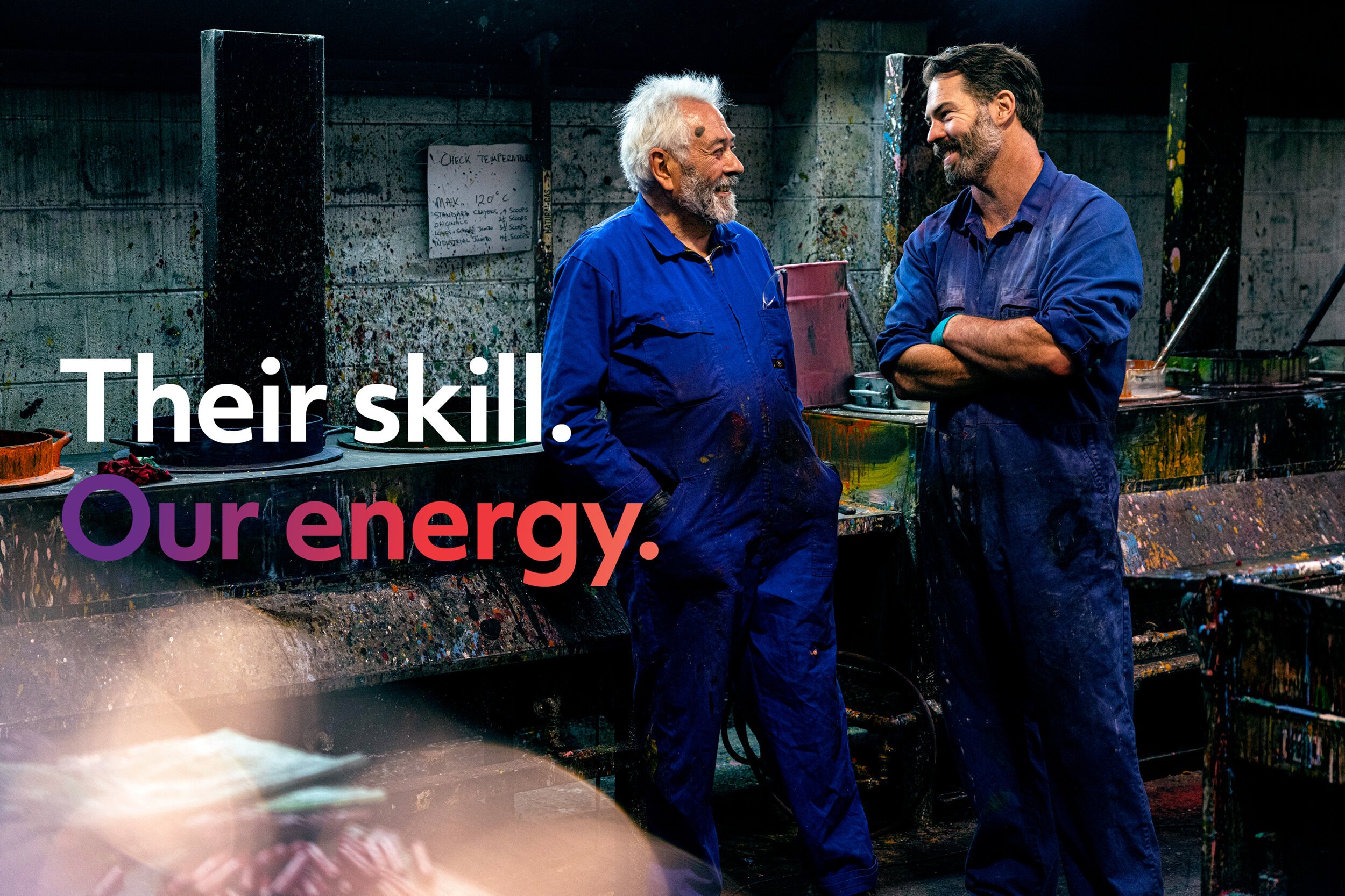 Two men in blue work uniforms, one older with white hair and the other younger with dark hair, smiling and talking in an industrial or artistic space with splattered paint and a whiteboard in the background.