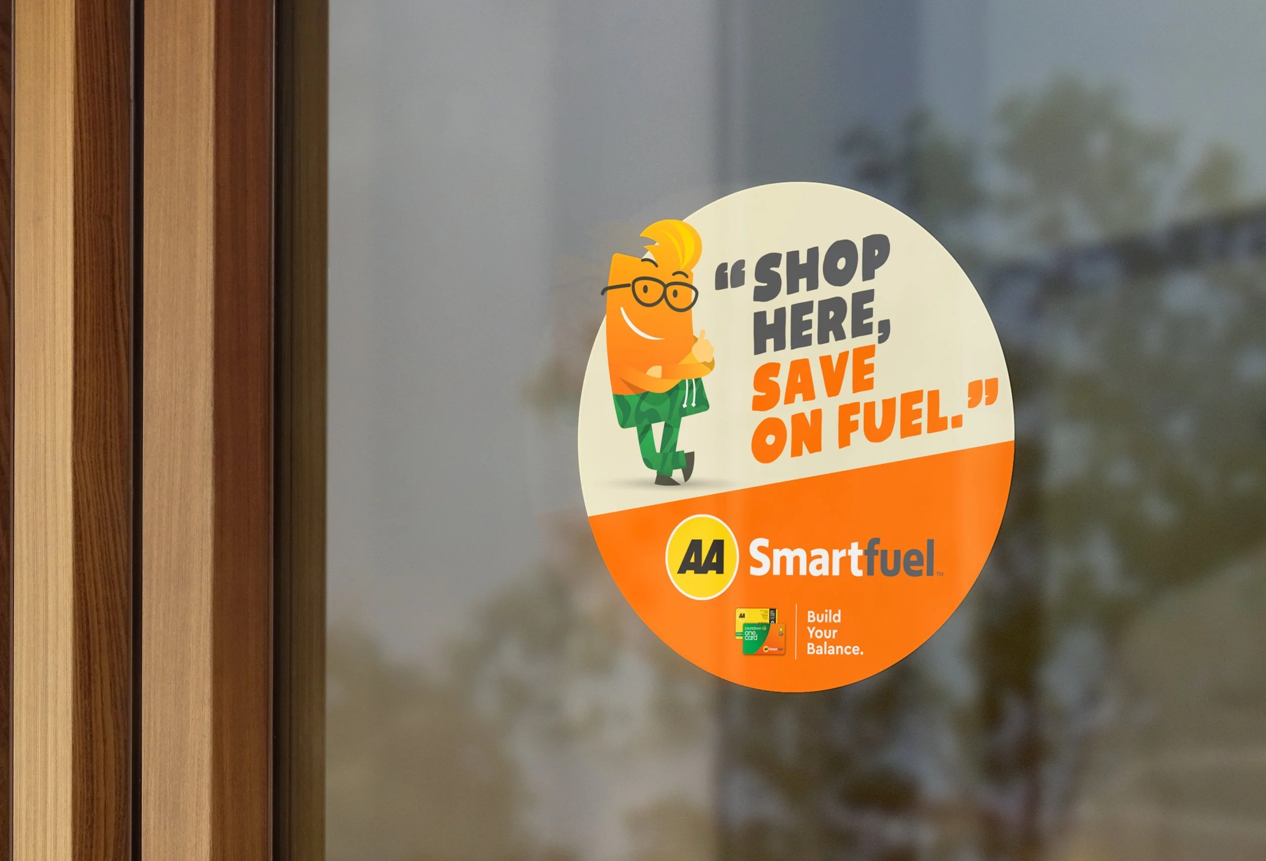 A circular sticker on a glass door or window features a cartoon orange character with glasses and green shorts, giving a thumbs-up. The sticker displays the message 'Shop here, save on fuel' and the logo 'AA Smartfuel' with the tagline 'Build Your Balance'.