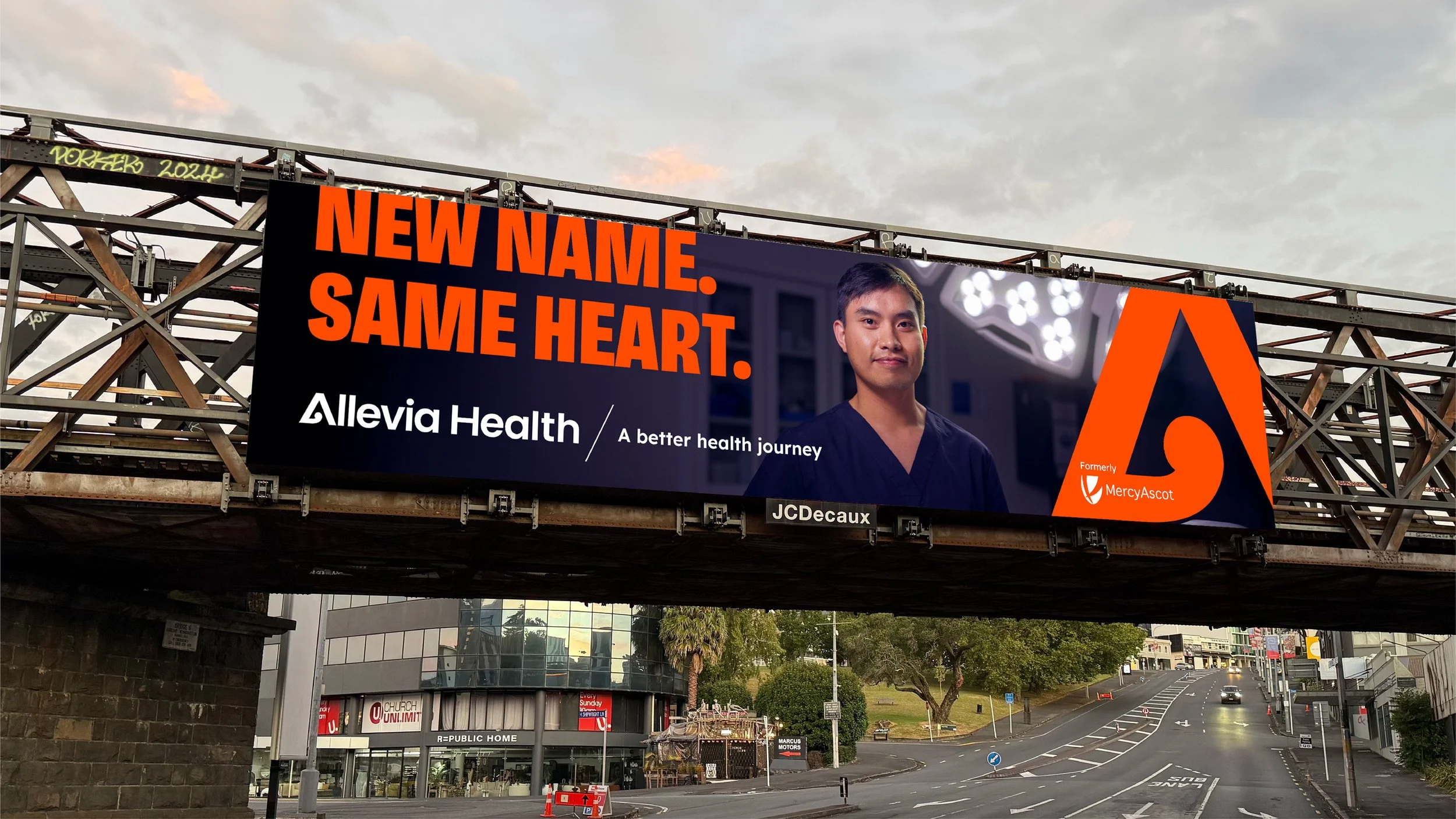 A billboard promoting Allevia Health with the slogan 'New Name. Same Heart.' features a young man in scrubs and the logos of MercyAscot. The billboard is located above an underpass on a city street observed at sunset.
