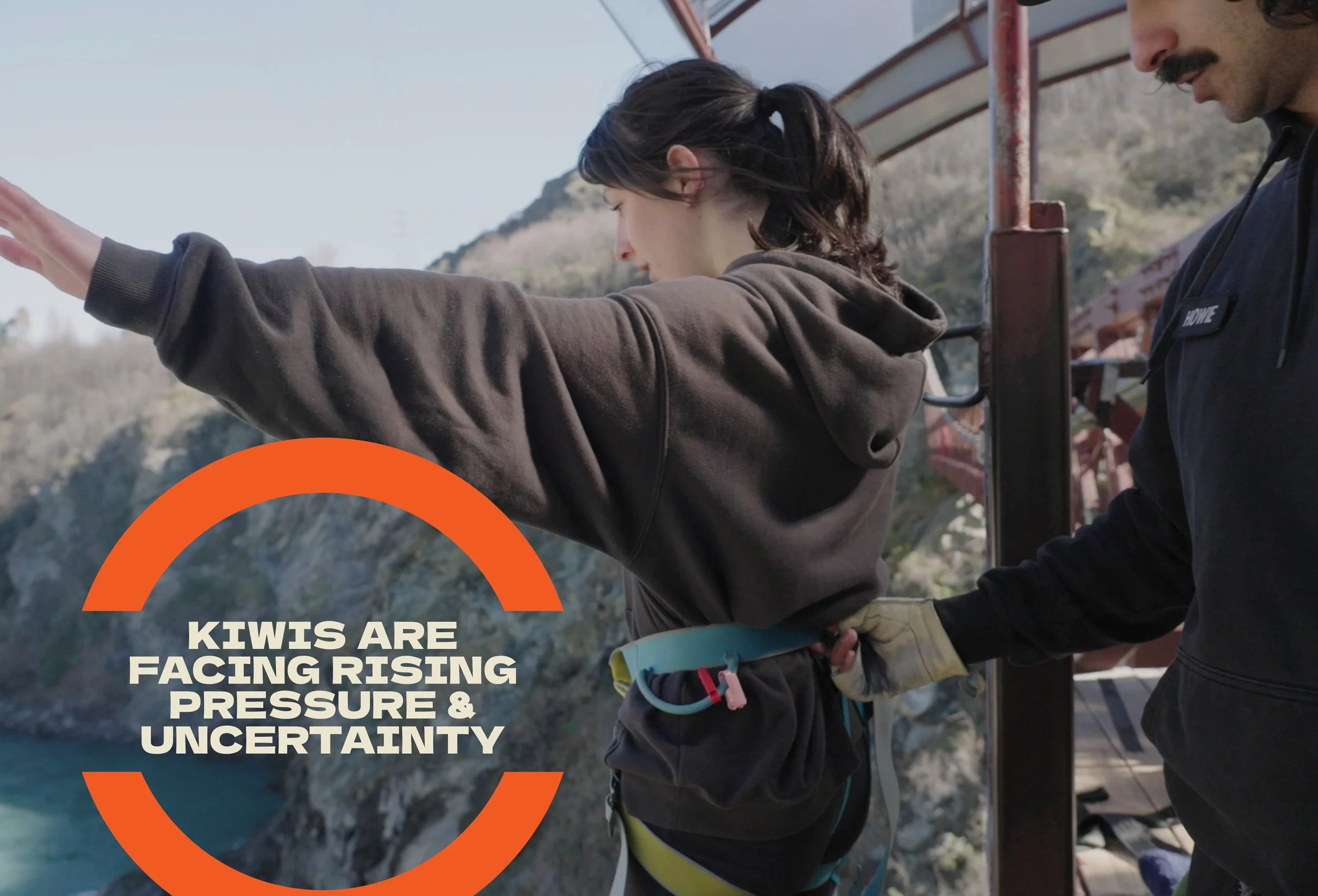 Two people, a woman and a man, are standing on a bridge overlooking a rocky canyon with water below. The woman is reaching out with her arm extended, and the man is looking at her, both focused on the view or an object she is pointing to. The woman has dark hair tied back and is wearing a brown hoodie, while the man has a mustache, glasses, and is wearing a black jacket with a name tag.