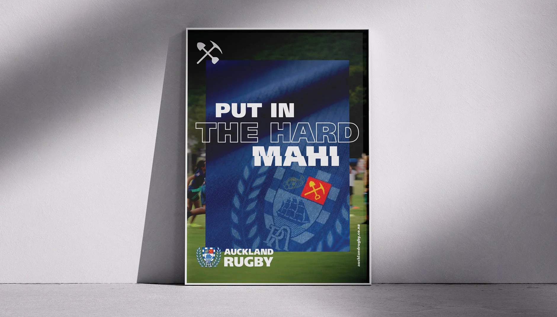A poster for Auckland Rugby features the words 'PUT IN THE HARD MAHI' with a rugby emblem and a small icon of an axe and pickaxe, displayed on a white wall.