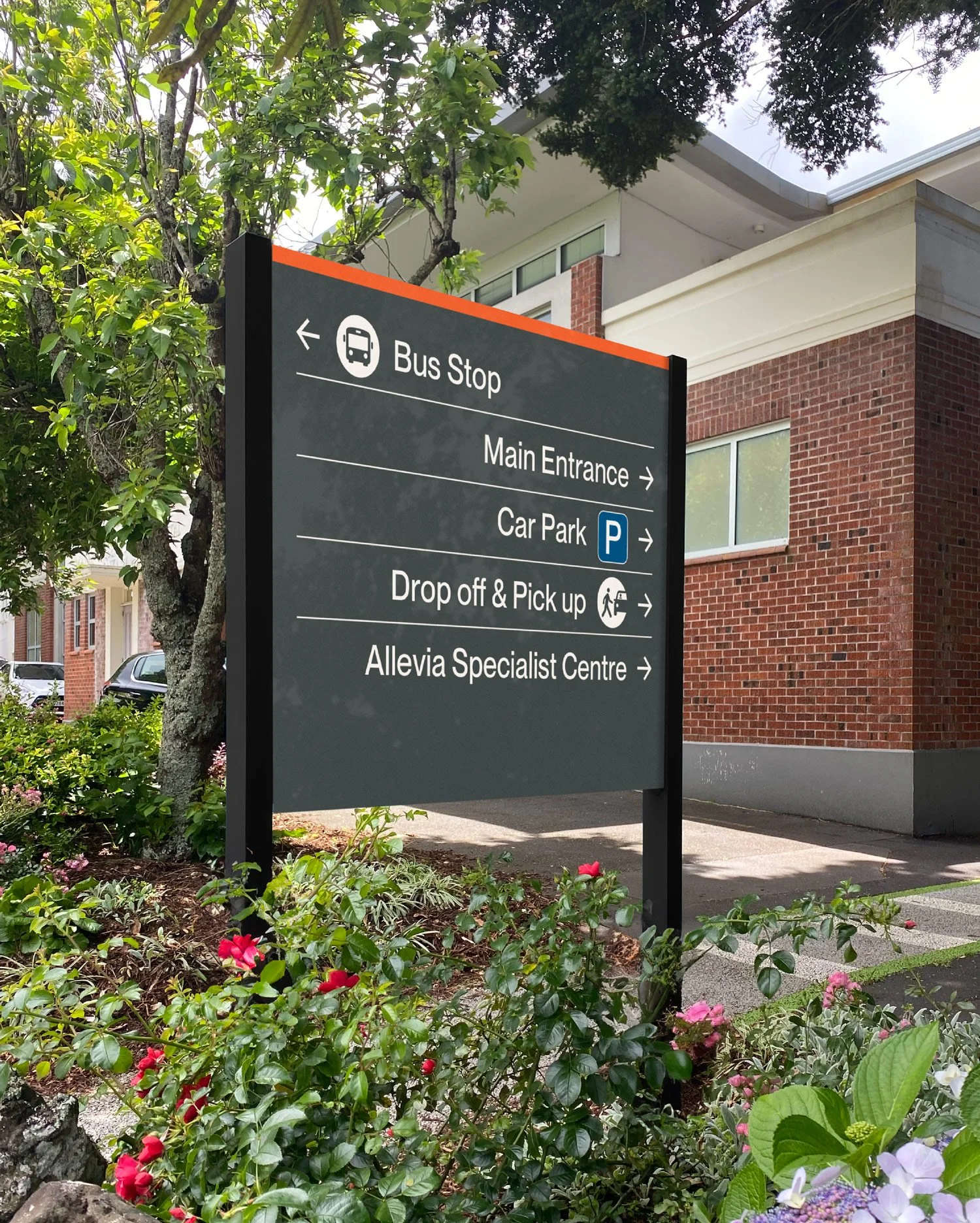 A large outdoor sign with directions for a bus stop, main entrance, car park, drop off and pick up, and a specialist center. The sign is surrounded by flowering plants and trees, with a brick building in the background.