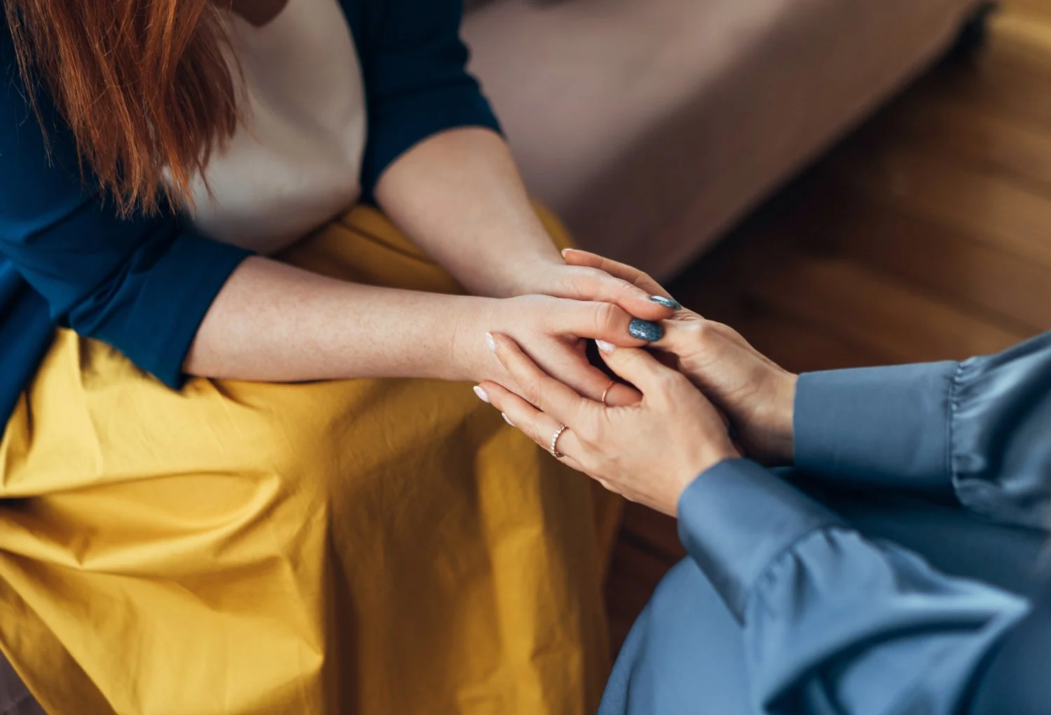 Two women are holding hands, one seated wearing a yellow skirt and the other standing or kneeling wearing a blue-gray long-sleeved shirt.