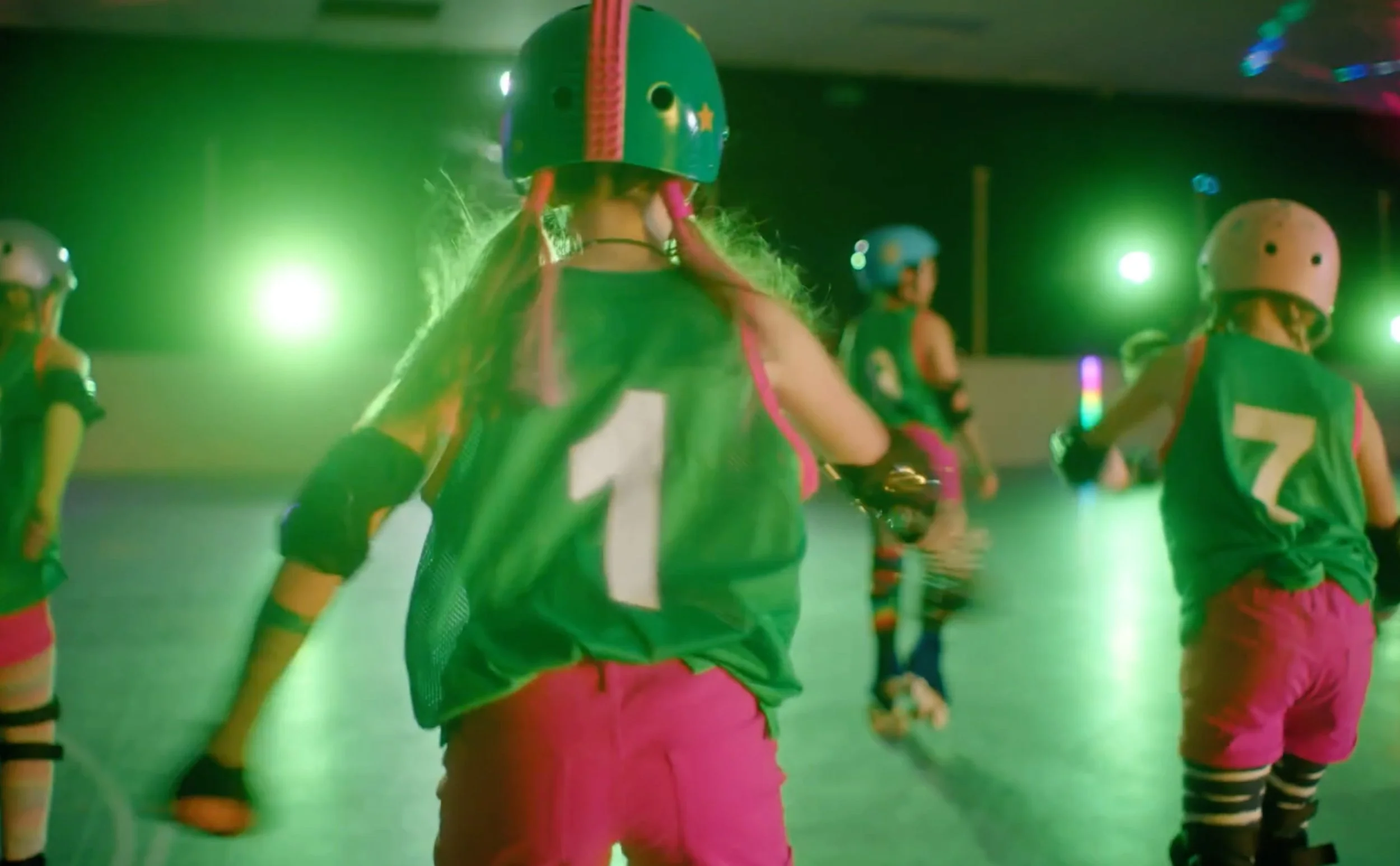 A group of young girls roller skating indoors under colorful green lighting. They are wearing helmets, elbow and knee pads, and athletic clothing, with some girls facing away from the camera.