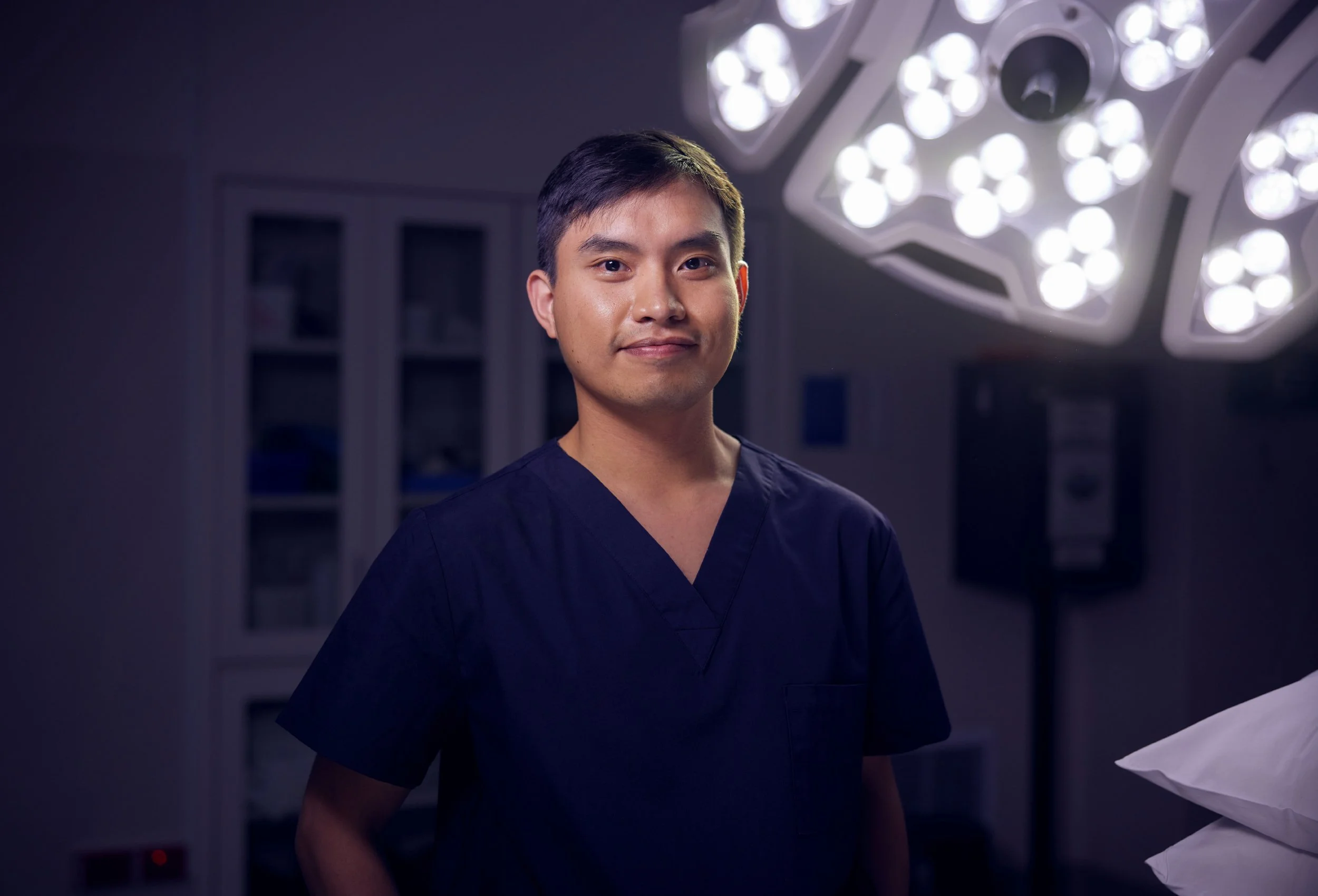 A young male nurse or medical professional in navy scrubs standing in a clinical setting with surgical lights overhead.