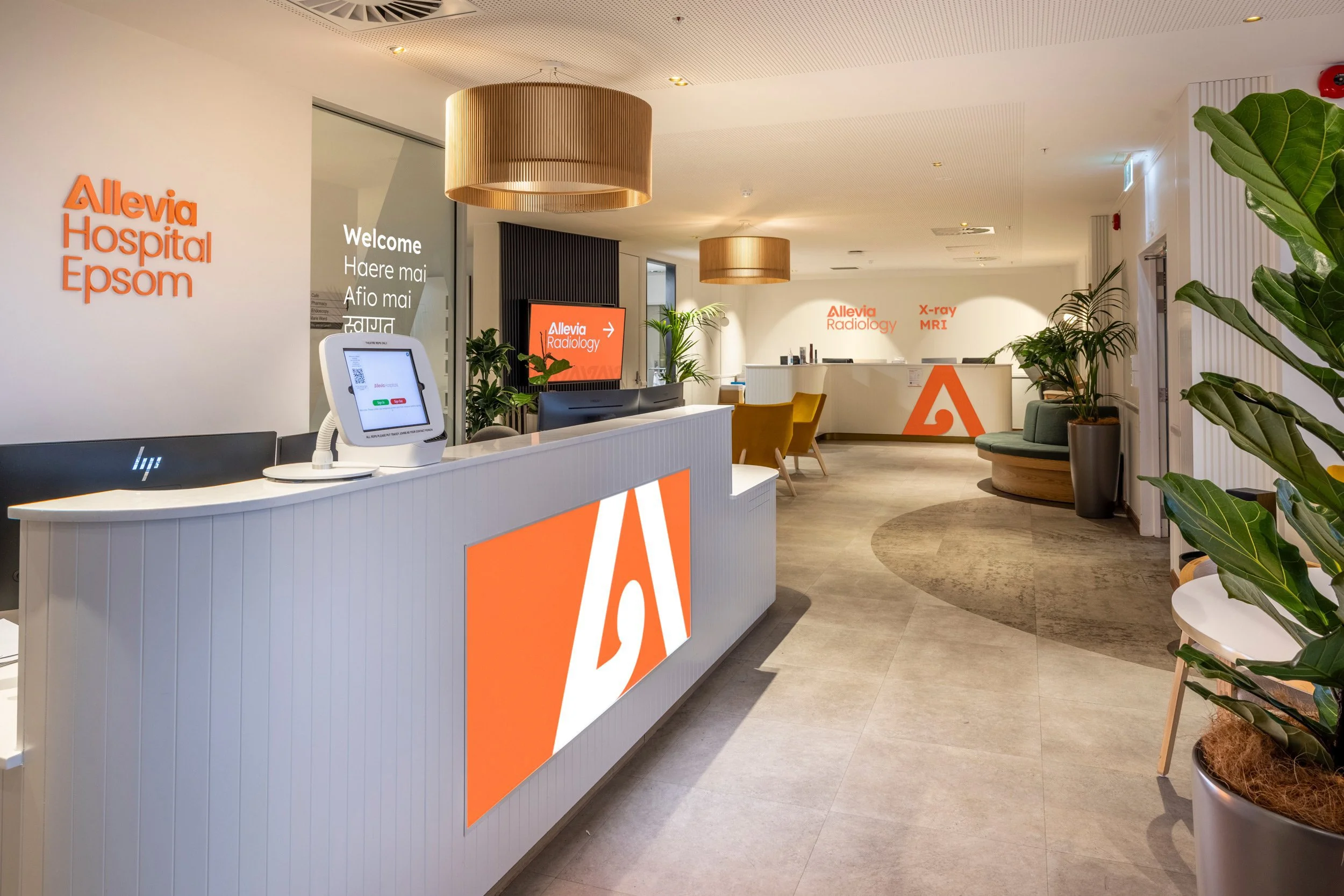 Reception area at Allevia Hospital Epsom with a white counter, plants, and signage indicating radiology services like X-ray and MRI, featuring modern lighting and decor.