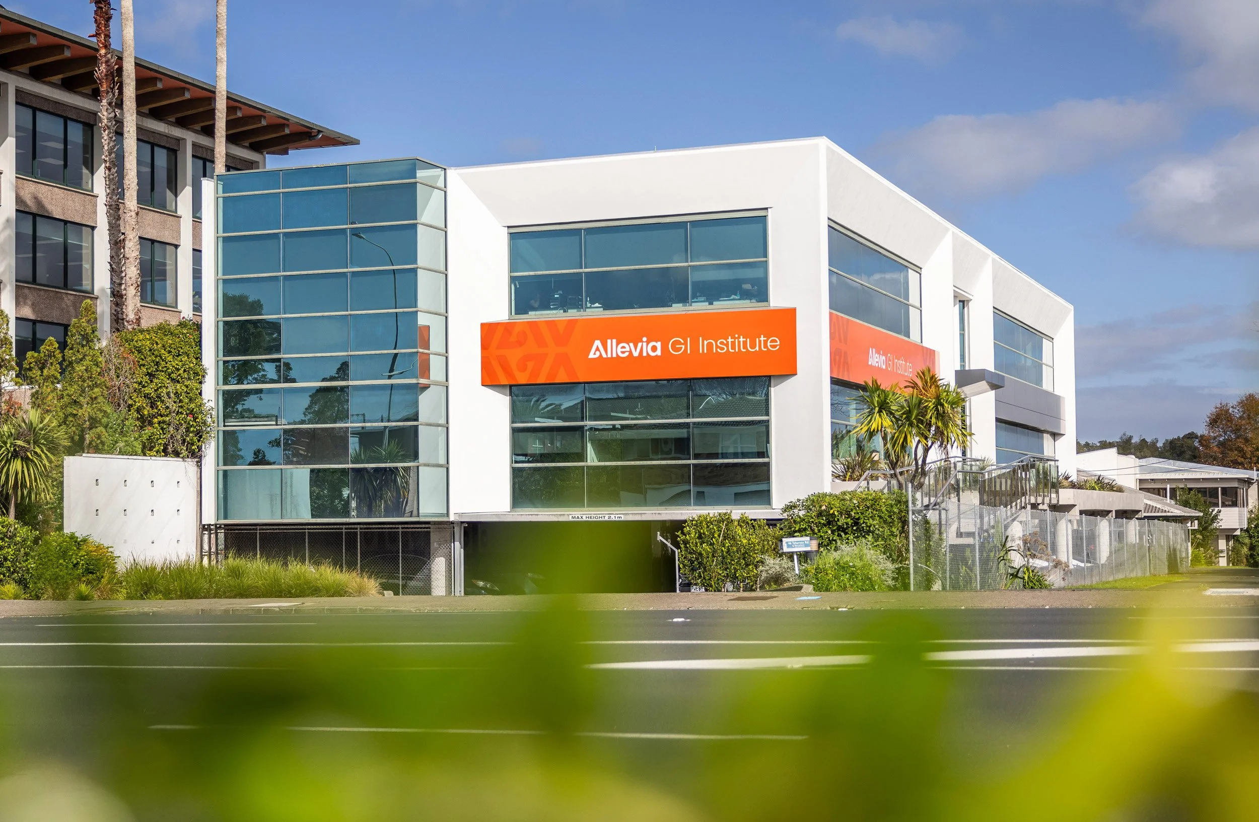 Modern multi-story building with large glass windows and a prominent orange sign reading 'Allevia GI Institute'; surrounded by greenery and trees, under a partly cloudy sky.