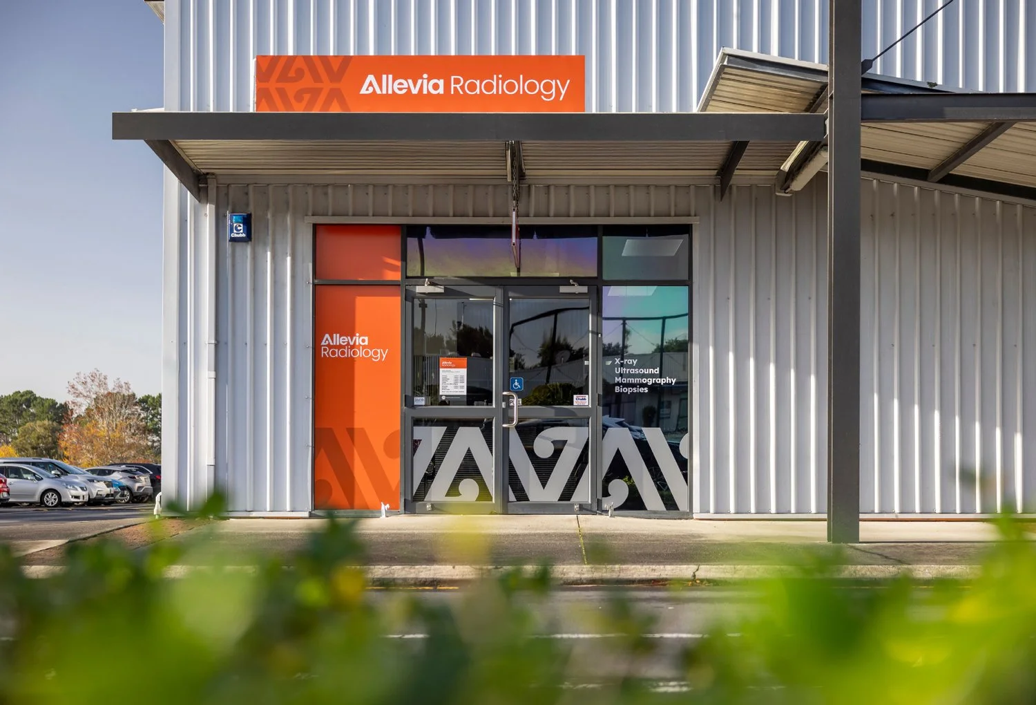 Exterior view of a medical imaging clinic named Allevia Radiology, with a glass door and a parking lot visible in the background.