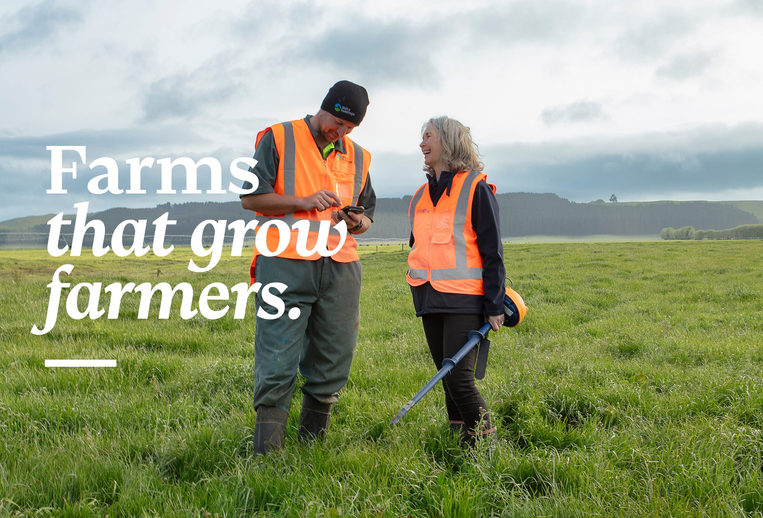 Two farmers in orange vests talking in a green field, one holding a metal detector, with rolling hills and cloudy sky in the background.