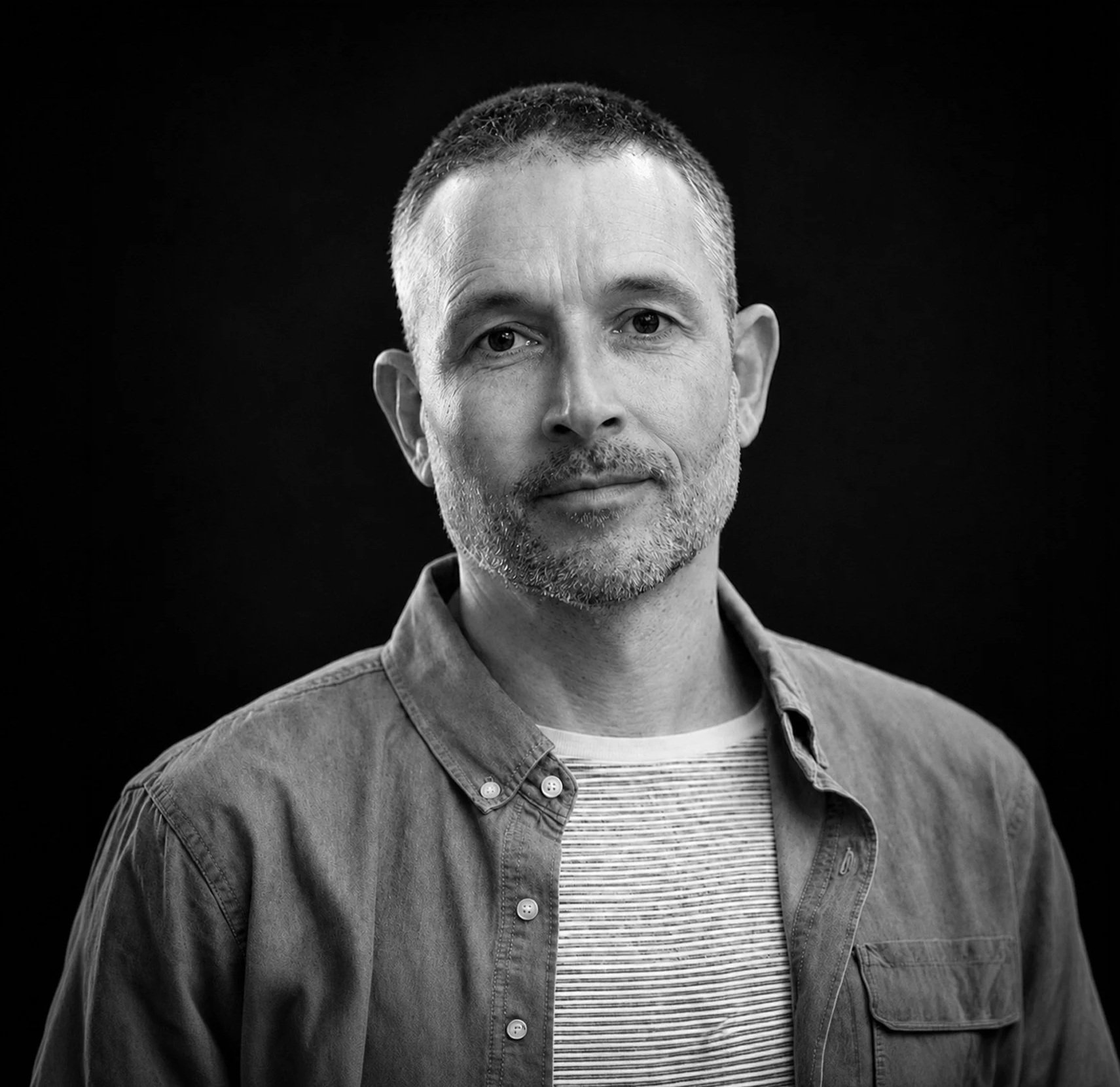 Black and white portrait of a middle-aged man with short hair, stubbled beard, wearing a button-up shirt over a striped t-shirt, standing against a dark background.