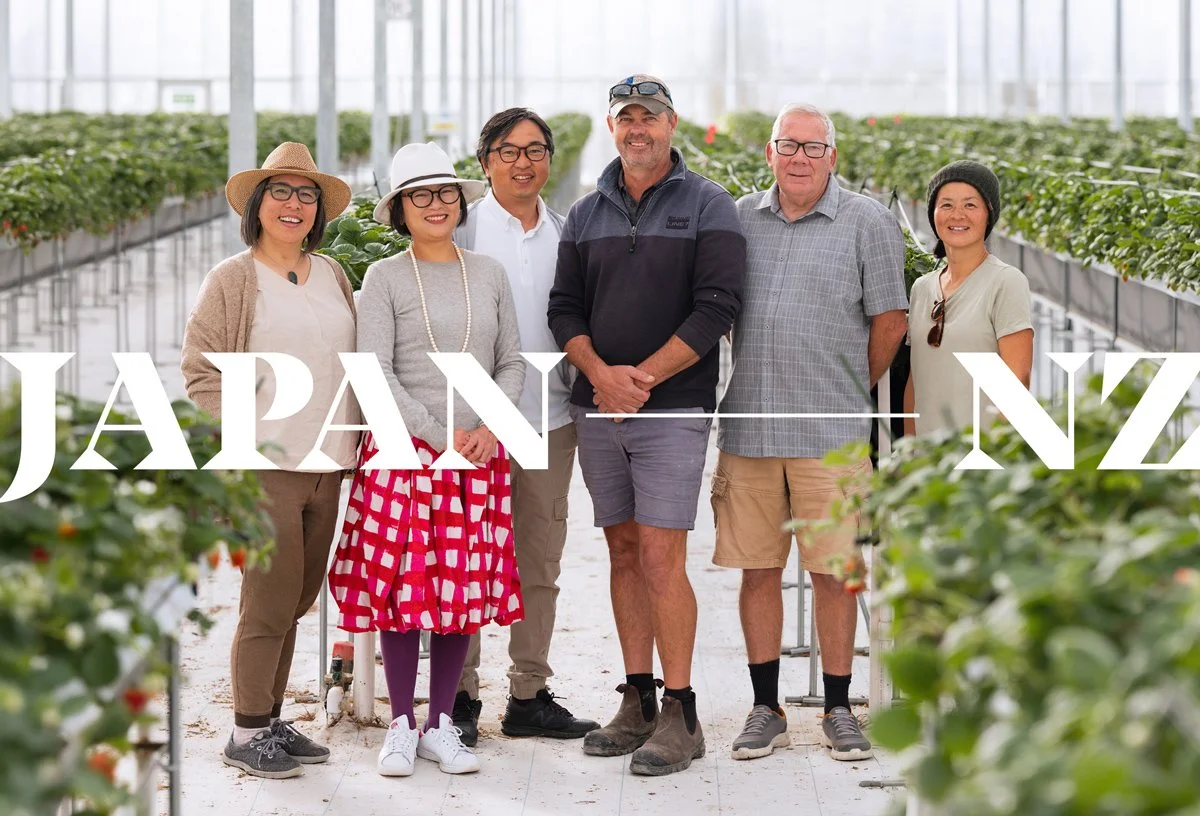 Group of six people standing inside a greenhouse with rows of strawberry plants, smiling at the camera, with the text 'JAPAN NZ' overlaid.