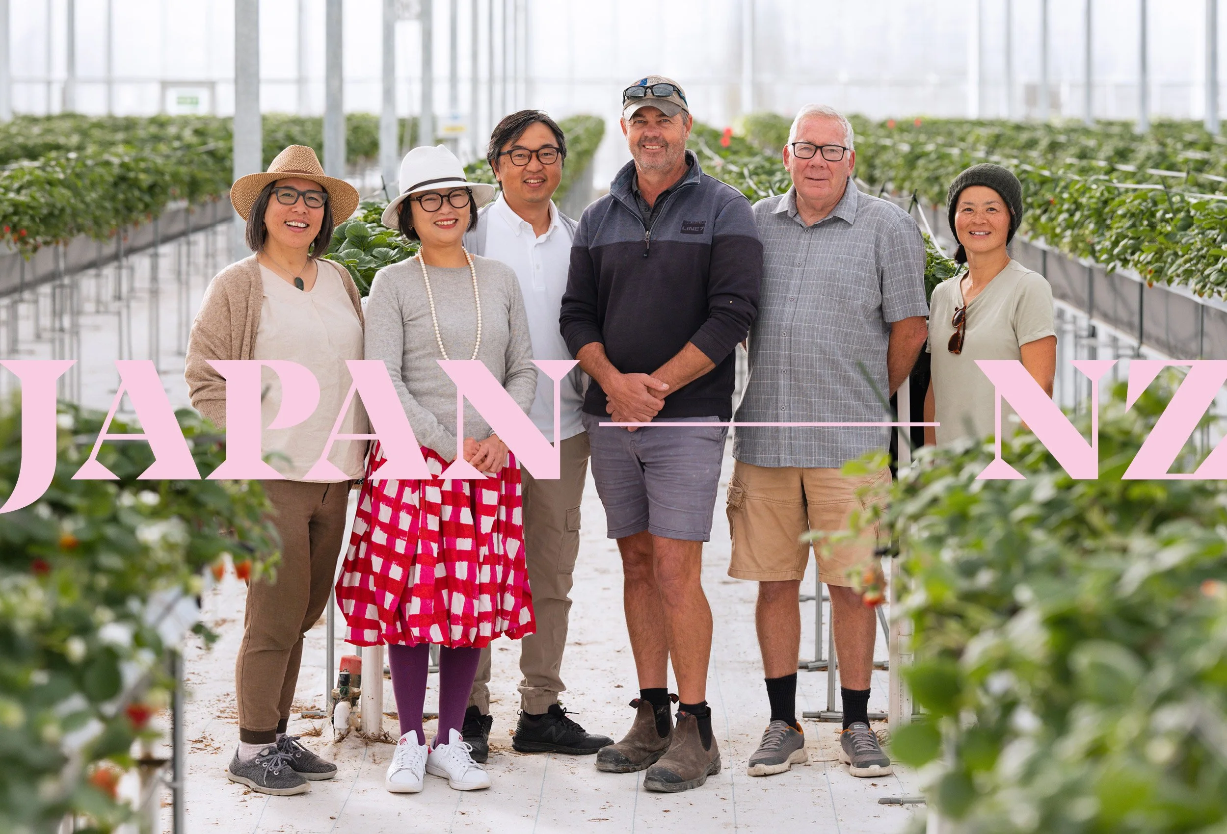A diverse group of people standing in a greenhouse with rows of strawberry plants, smiling at the camera.