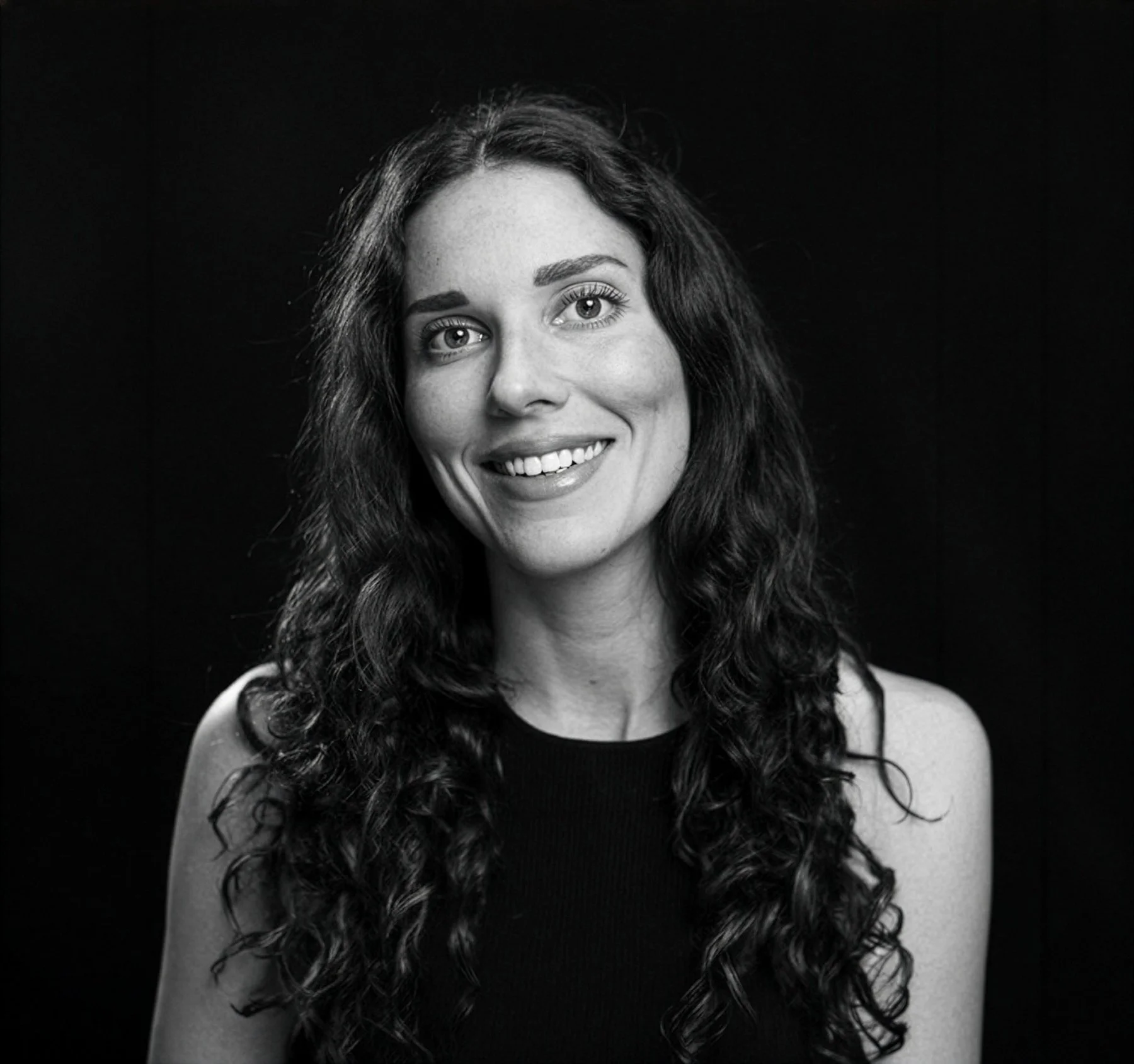 A black-and-white portrait of a smiling woman with long, curly hair, wearing a sleeveless top, against a dark background.