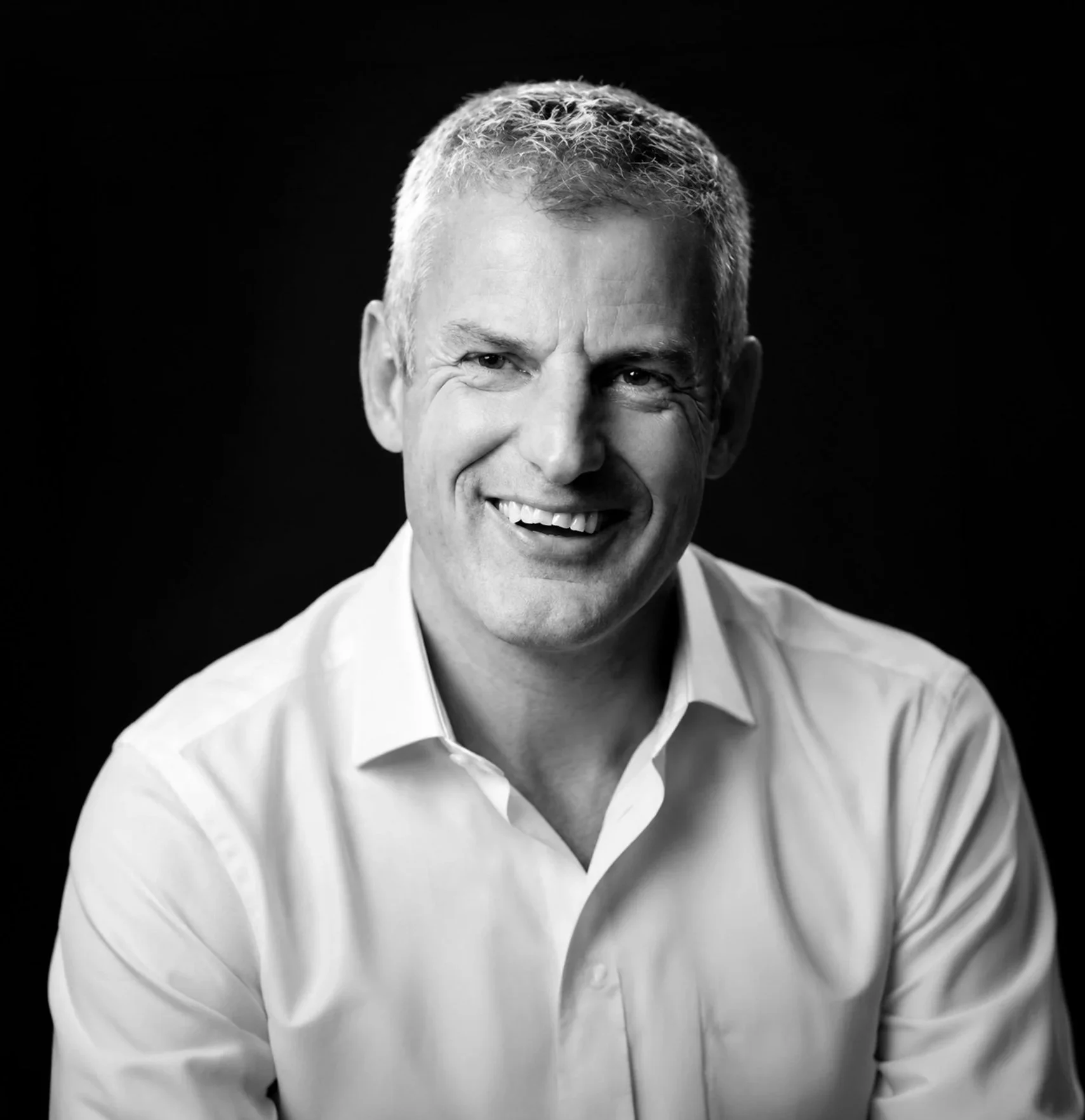 Black and white portrait of smiling man with short light-colored hair, wearing a white collared shirt, against a dark background.