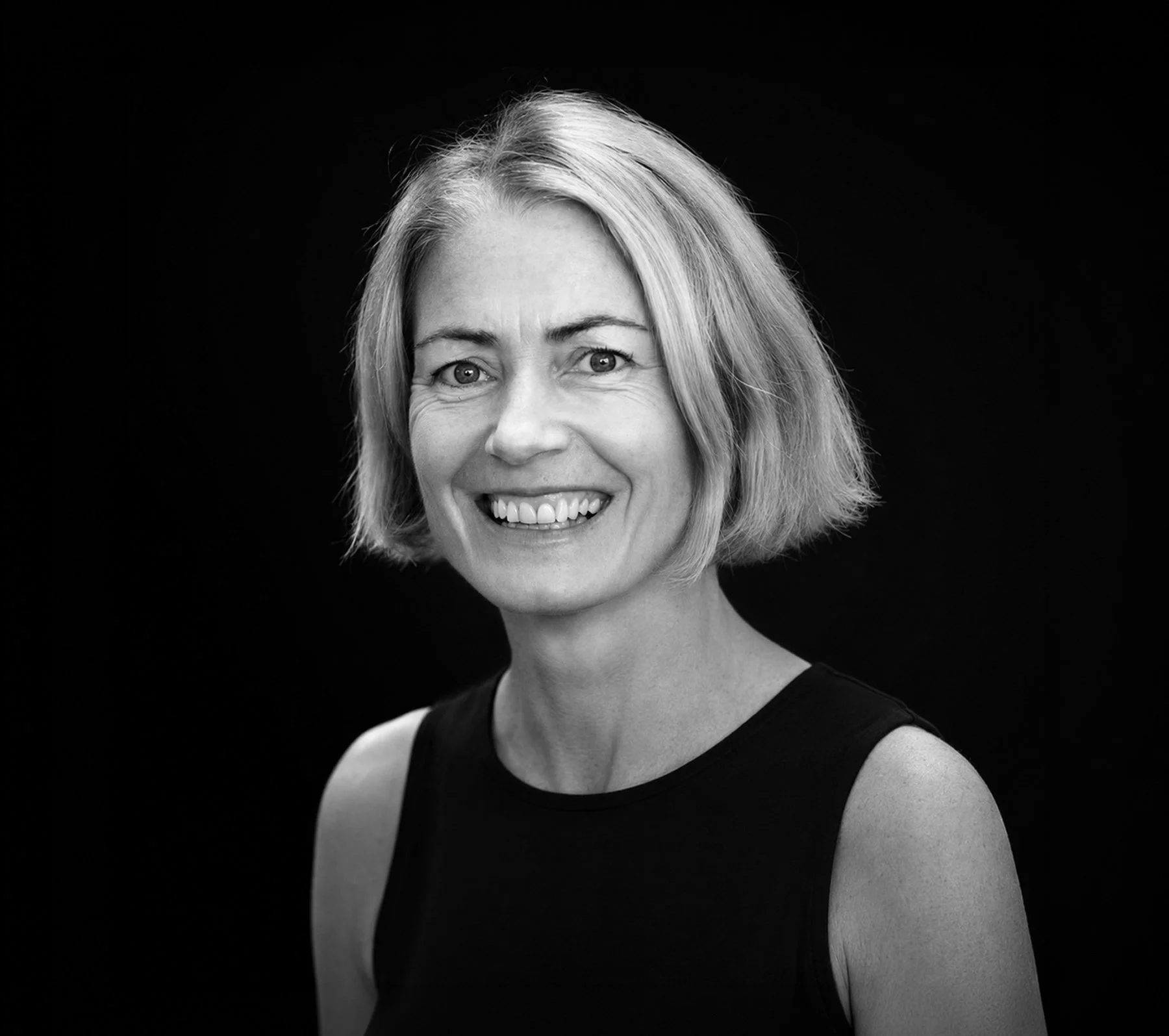 Black and white photo of a woman with short blonde hair, smiling, wearing a sleeveless black top, against a black background.