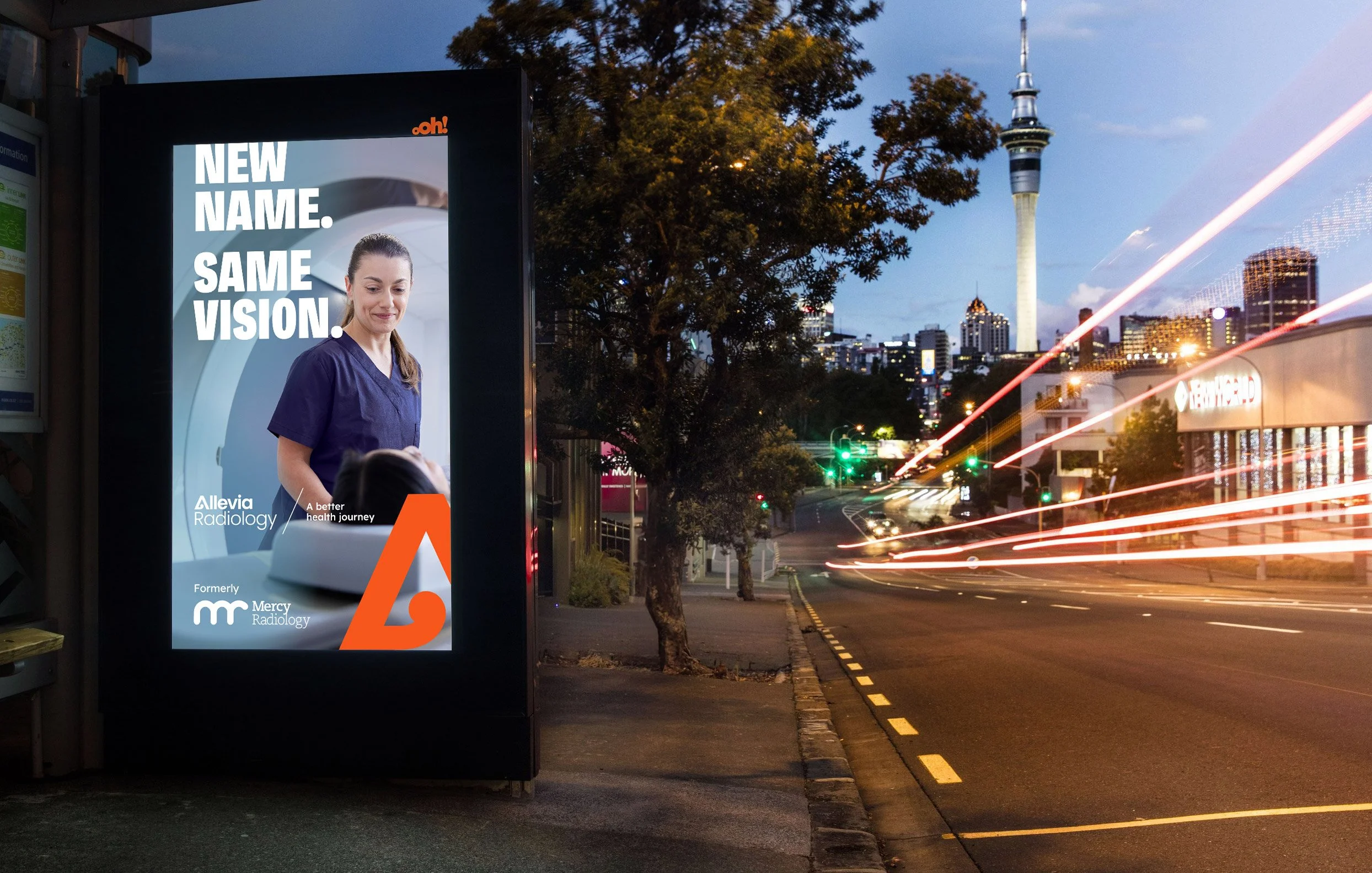 An electronic billboard advertising Allevia Radiology with the slogan 'New Name. Same Vision.' On a city street during dusk, with light trails from passing vehicles, a tall tower in the background, and a tree partially obscuring the billboard.