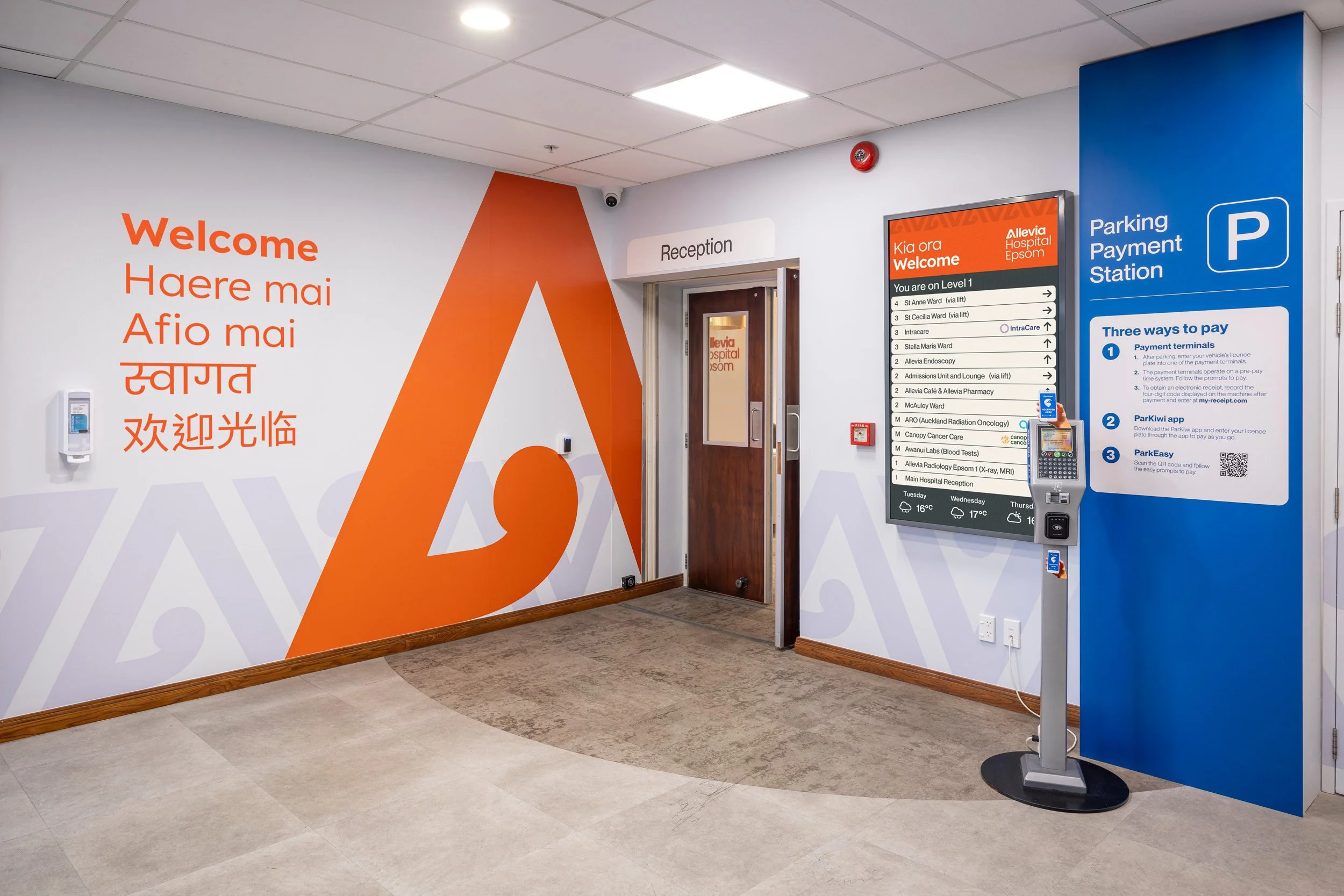 Hospital lobby with a large orange and white wall that says "Welcome" in multiple languages, a reception area with a sign, and a digital directory listing departments and their locations. There is a parking payment station on the right side and a blue wall with a parking sign.