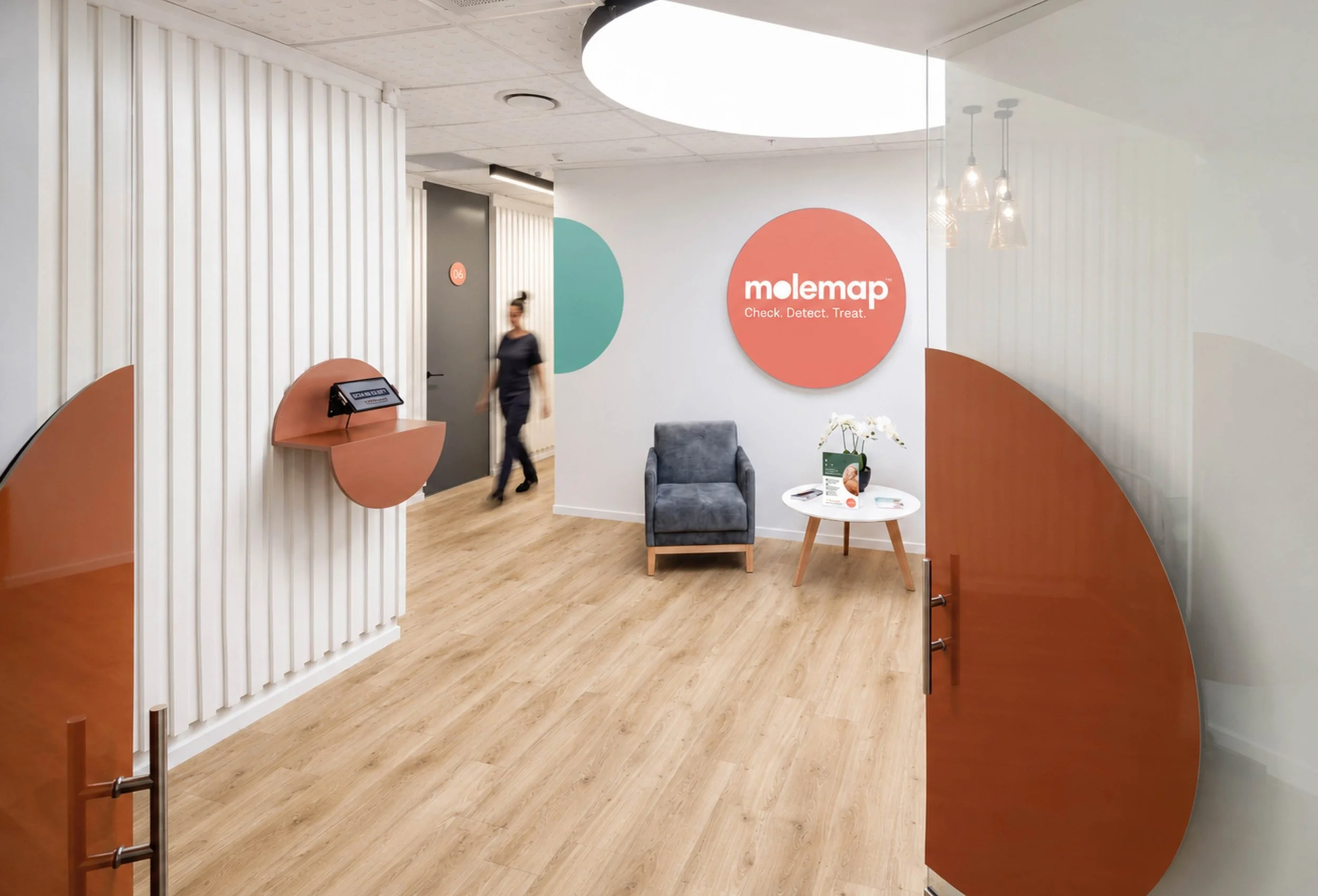 Modern medical office or clinic interior with a reception area, featuring a large circular light fixture on the ceiling, a small table with informational brochures, a comfortable gray armchair, and a woman in medical scrubs walking past a large round sign that reads 'molemap Check. Detect. Treat.' with white and pastel colored wall panels.