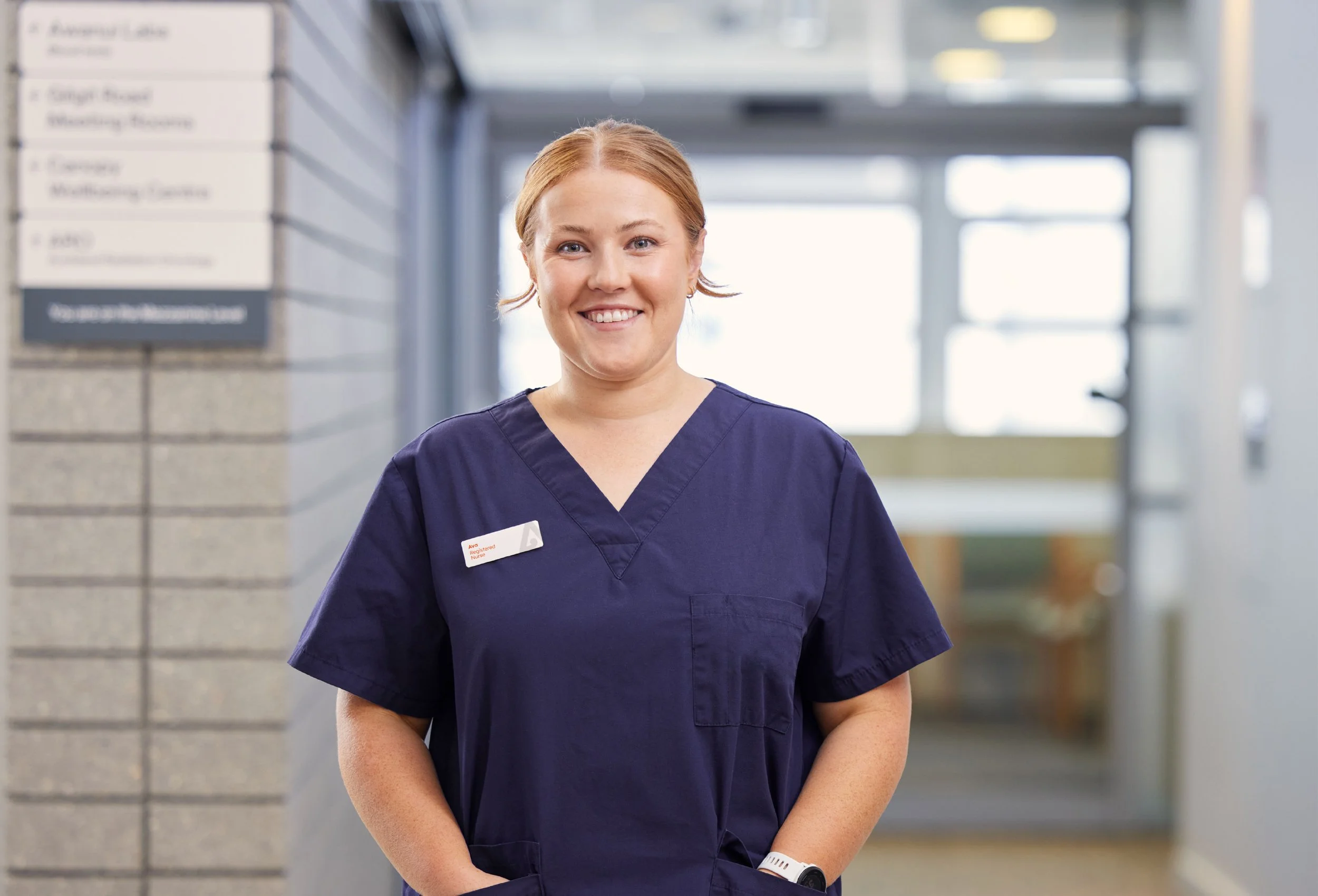 A smiling female healthcare worker standing in a hospital corridor, wearing navy scrubs and a white name badge, with a blurred background of hospital signage and windows.
