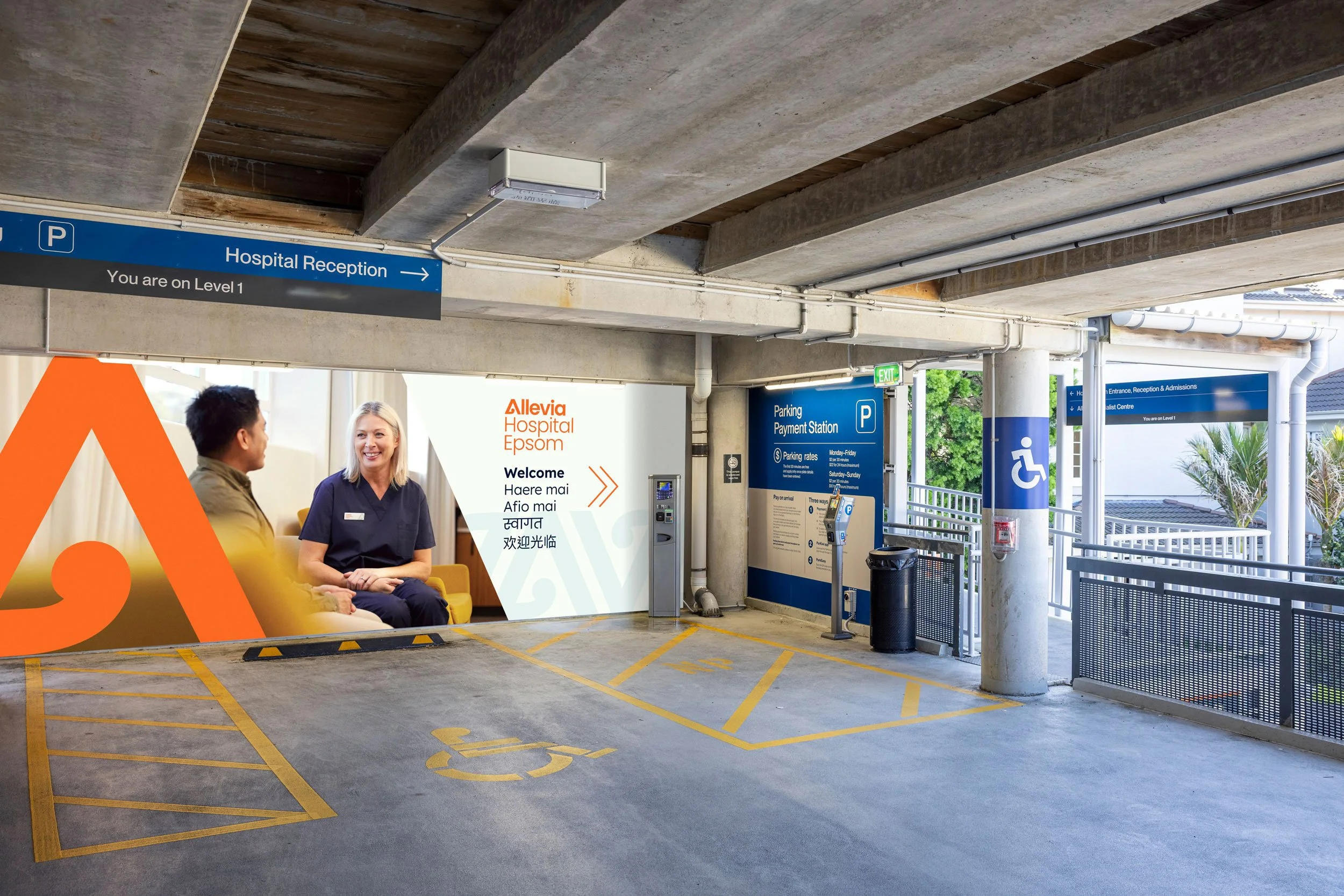 Underground parking garage with designated handicapped parking spaces and clear yellow markings. A parking payment station is visible near the entrance. Overhead signs direct visitors to the hospital reception on level 1, with additional signs indicating the hospital entrance, reception, admissions, specialist center, and an accessible restroom. There are two people, a man and a woman, sitting and conversing near the exits. The setting includes concrete beams and an open-air section with some greenery visible.
