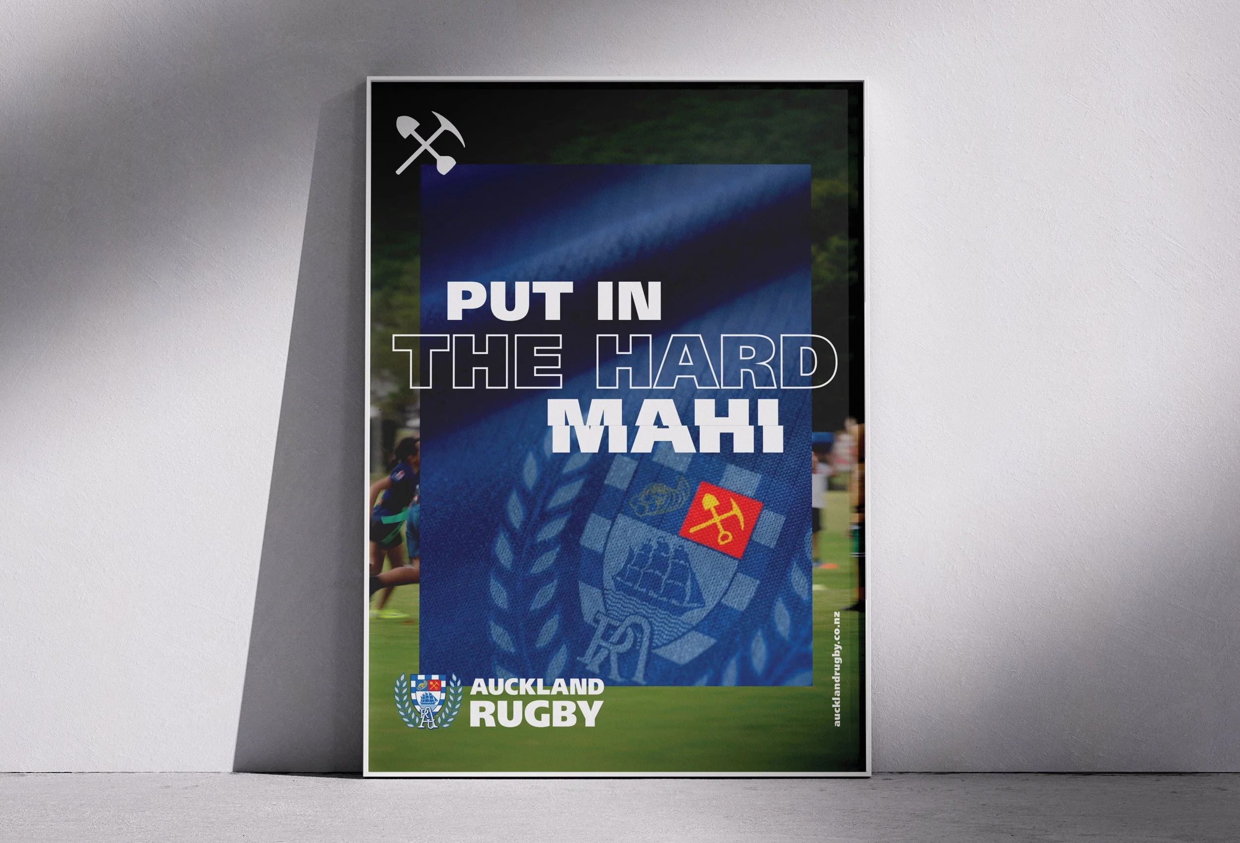 A poster on the wall promoting Auckland Rugby with the slogan 'Put in the Hard Mahi' and an icon of a pickaxe and hammer. The poster features rugby players in action and elements related to Auckland Rugby.