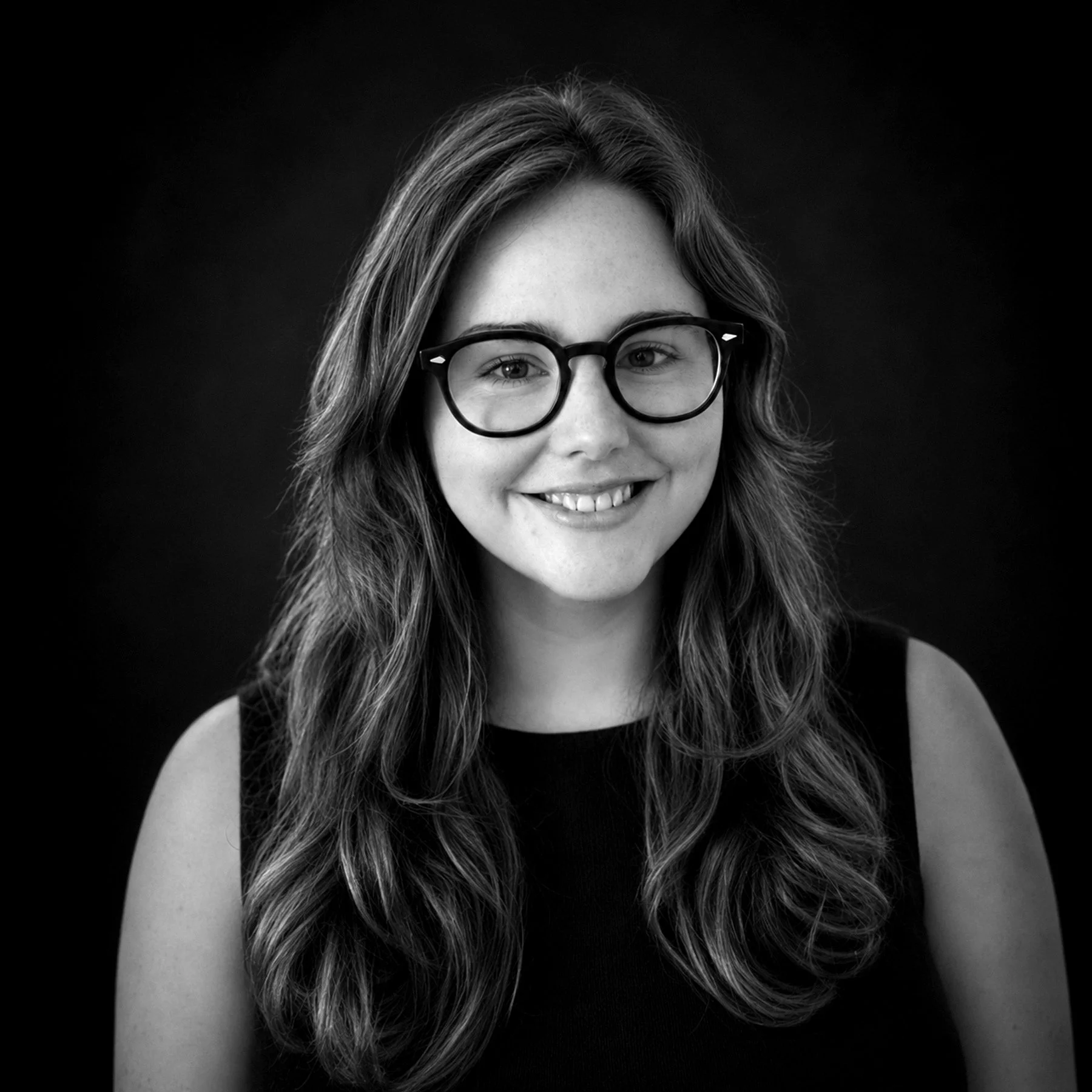 A black and white portrait of a young woman with wavy hair, wearing glasses, smiling at the camera.